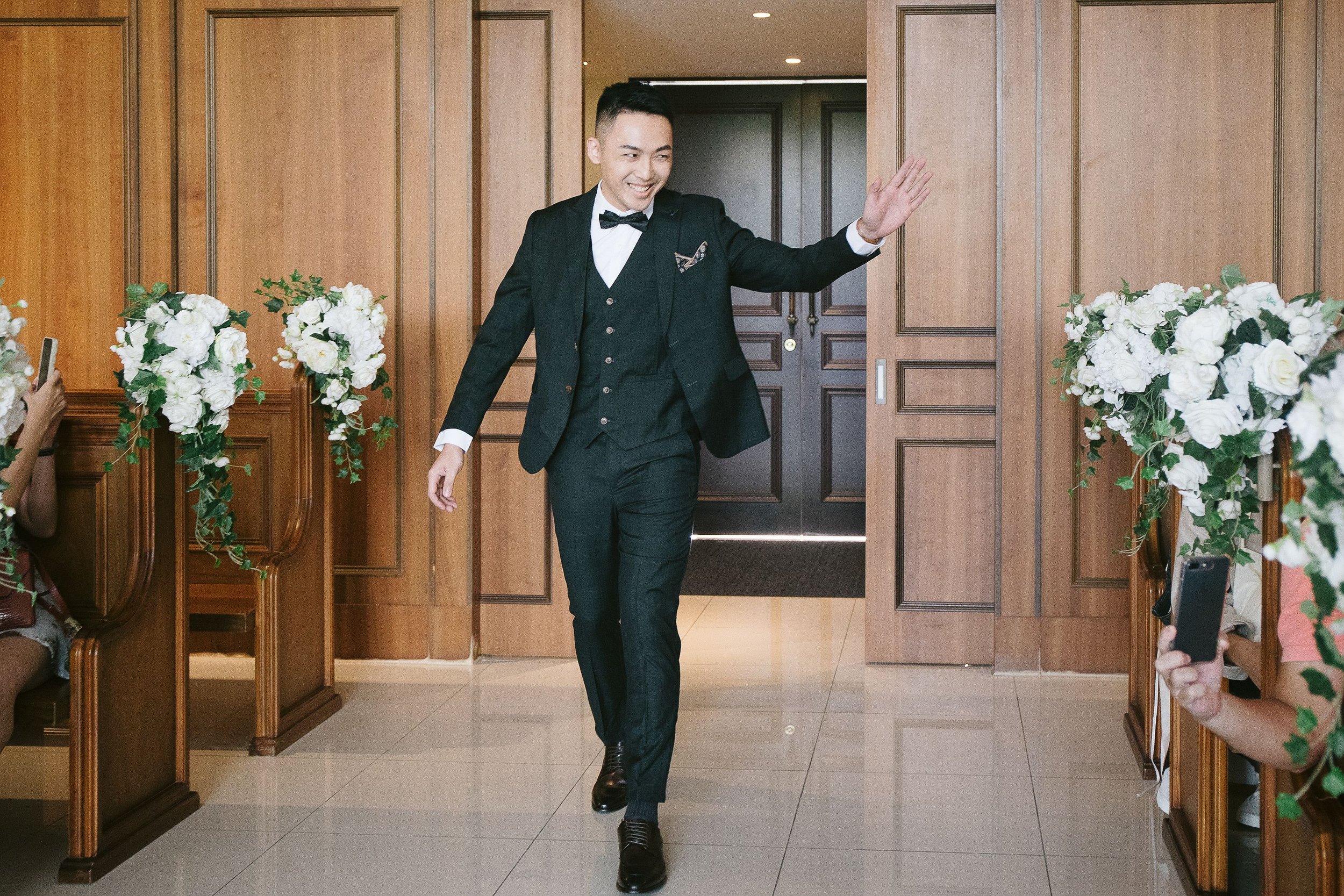 A young man in a black tuxedo and bow tie waving and smiling as he walks through a doorway decorated with white flowers for a special occasion, possibly a wedding.