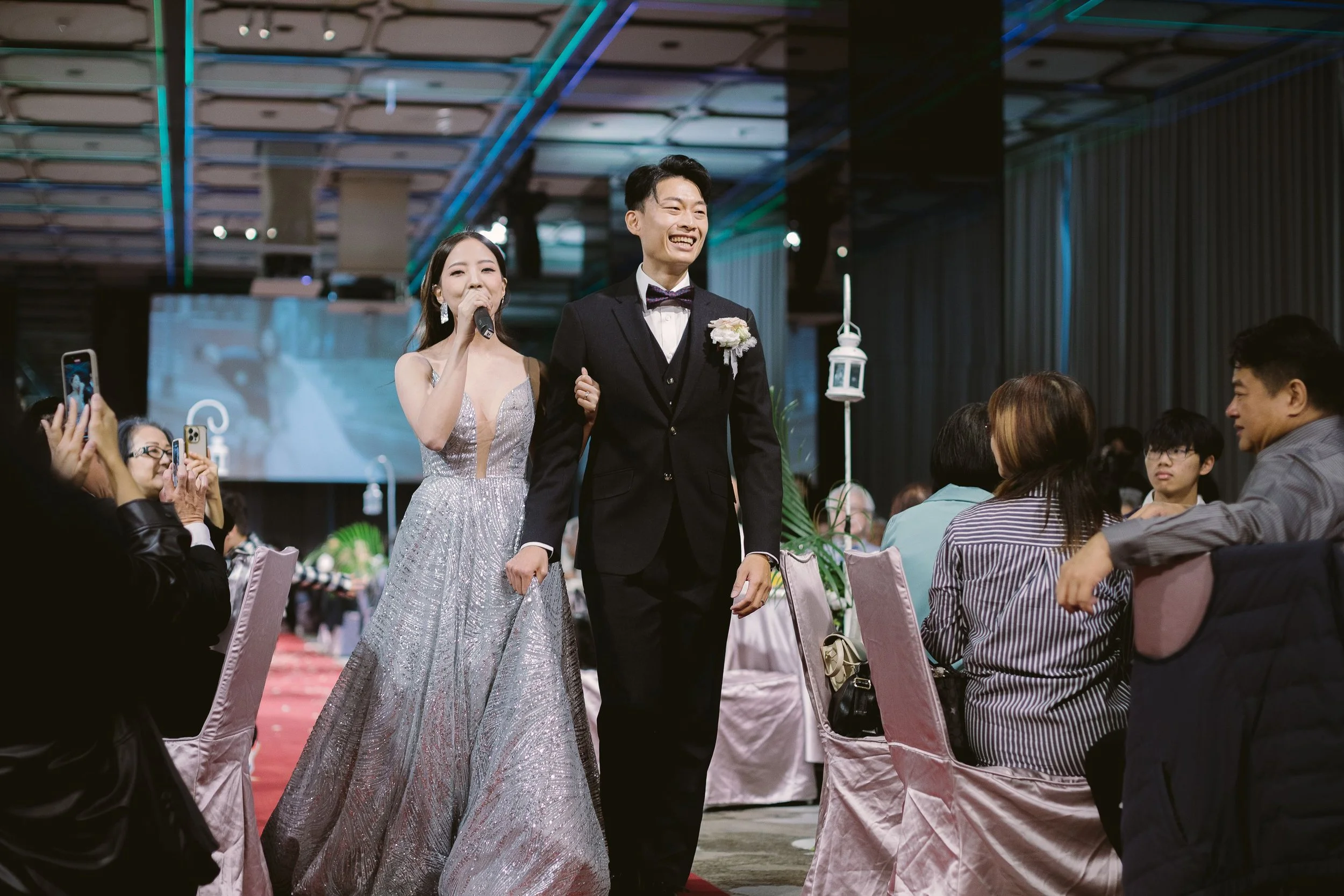 A bride and groom walking arm-in-arm at their wedding reception, smiling, with guests seated around them and taking photos.