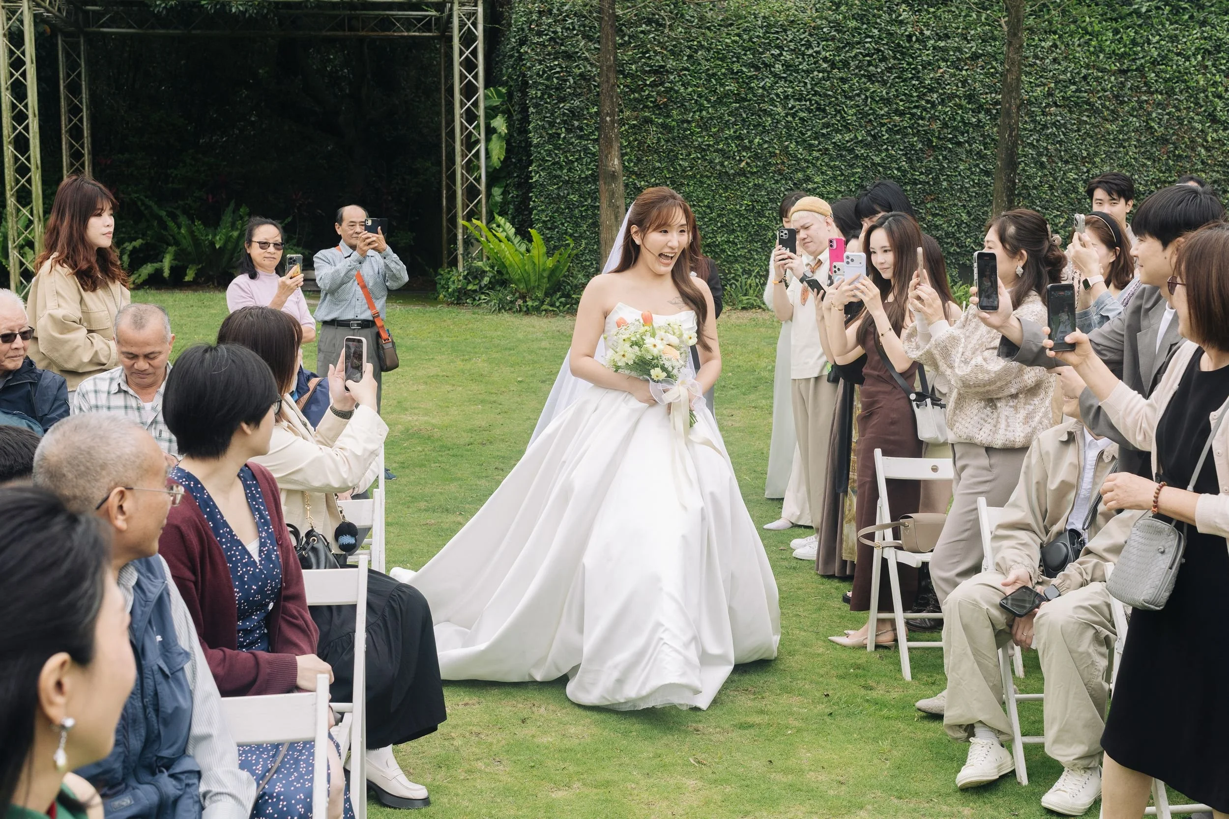 A bride in a white wedding dress holding a bouquet of flowers is smiling and walking down an outdoor aisle surrounded by seated guests and onlookers, many of whom are taking photos.