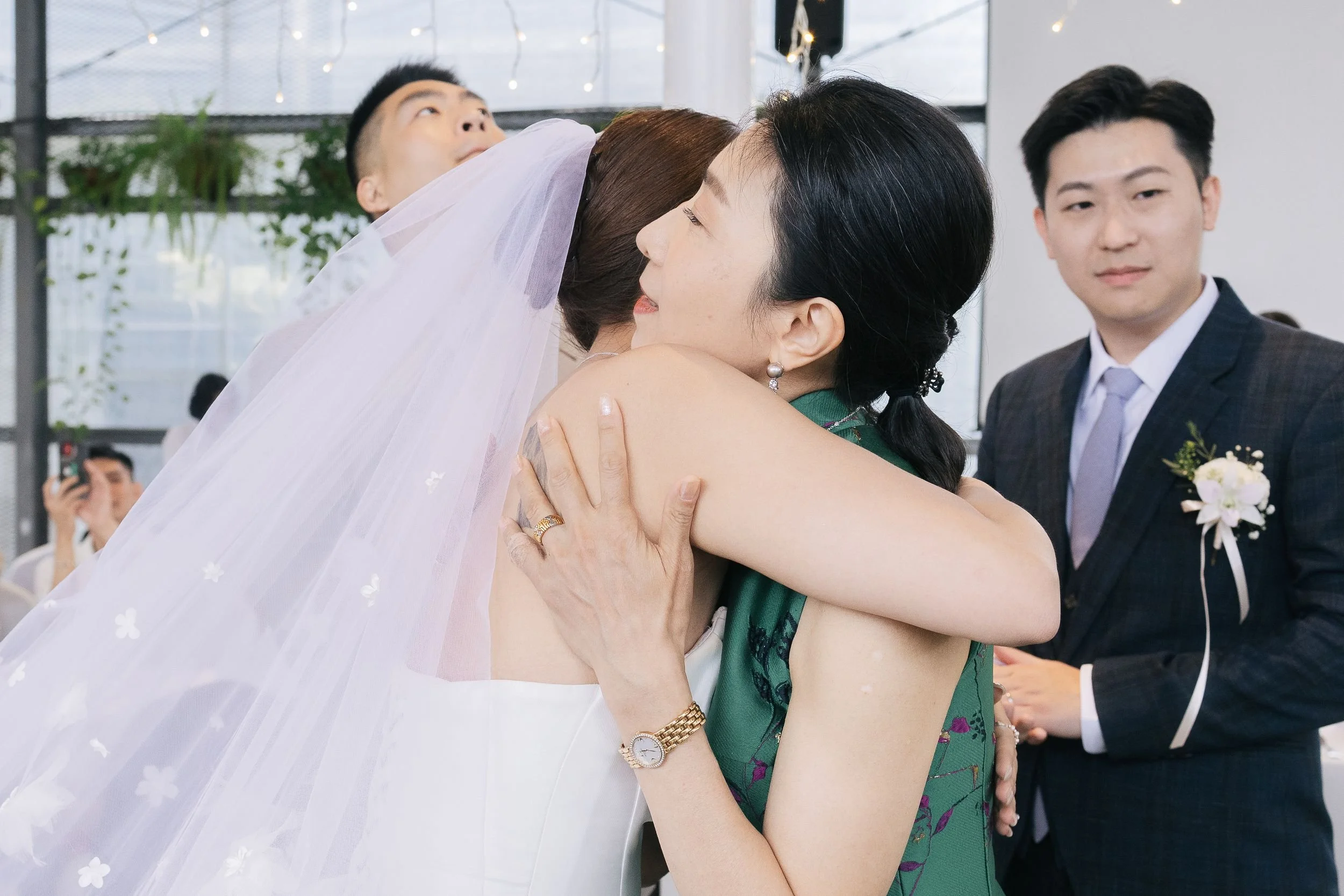 A woman in a green dress is hugging a bride who is wearing a white veil. A man in a suit is watching them, and another person is taking a photo in the background at a wedding celebration.