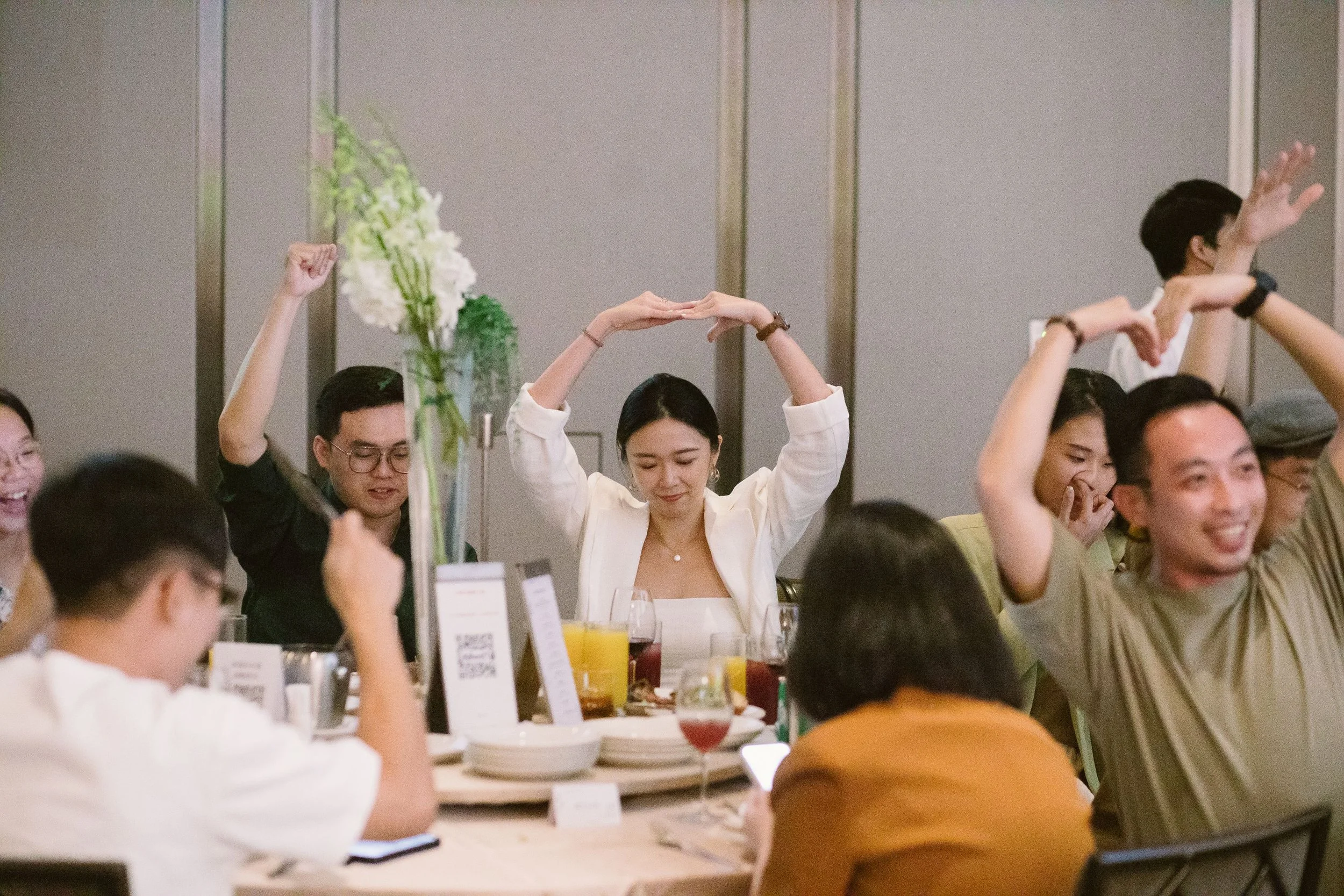 People sitting at a banquet table with their hands raised, some in prayer and others voting, with food and drinks on the table and floral arrangements in the background.