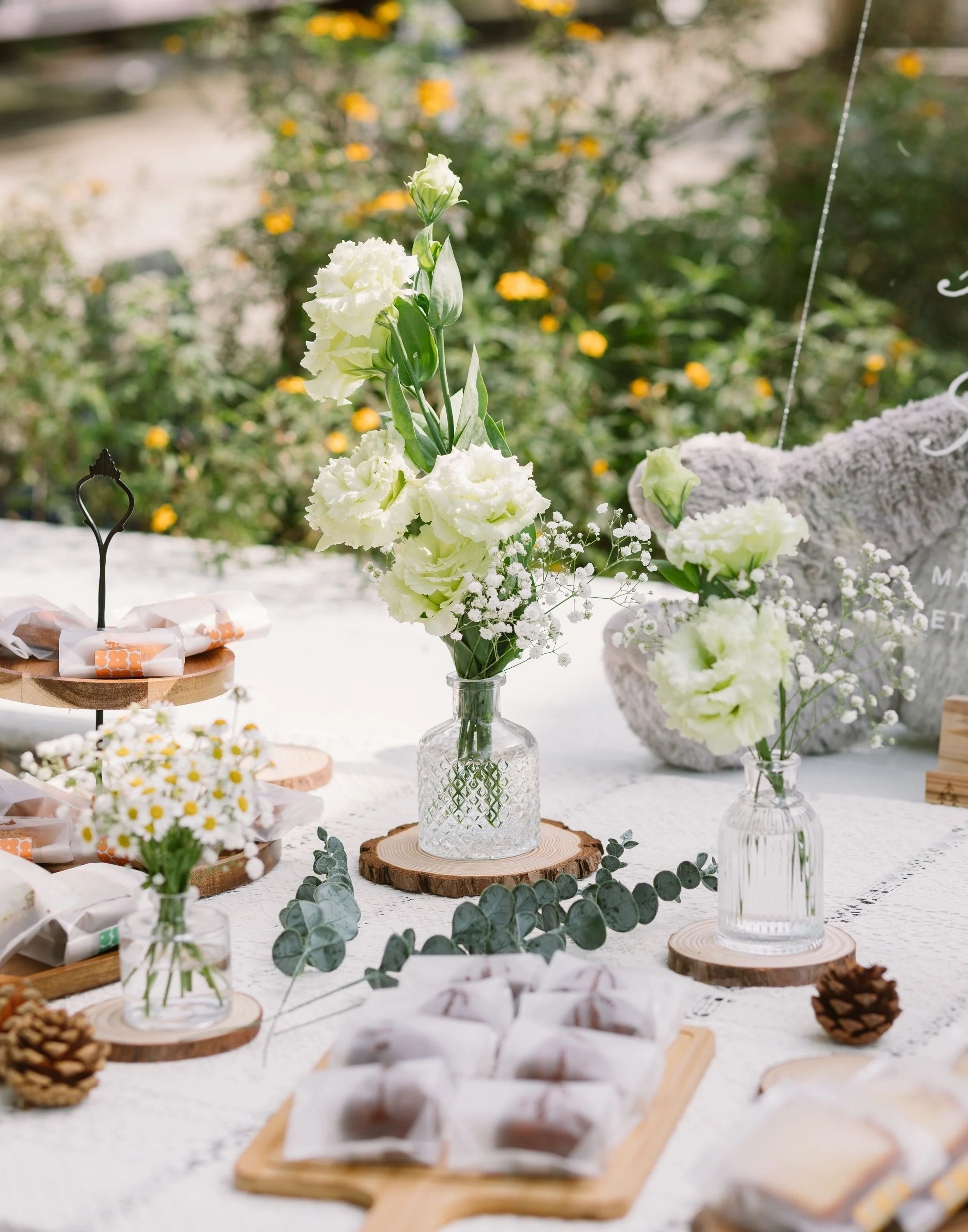 Decorative table with white flowers in glass vases, wooden slices, pine cones, and small wrapped items, outdoors with blurred green foliage and yellow flowers in the background.