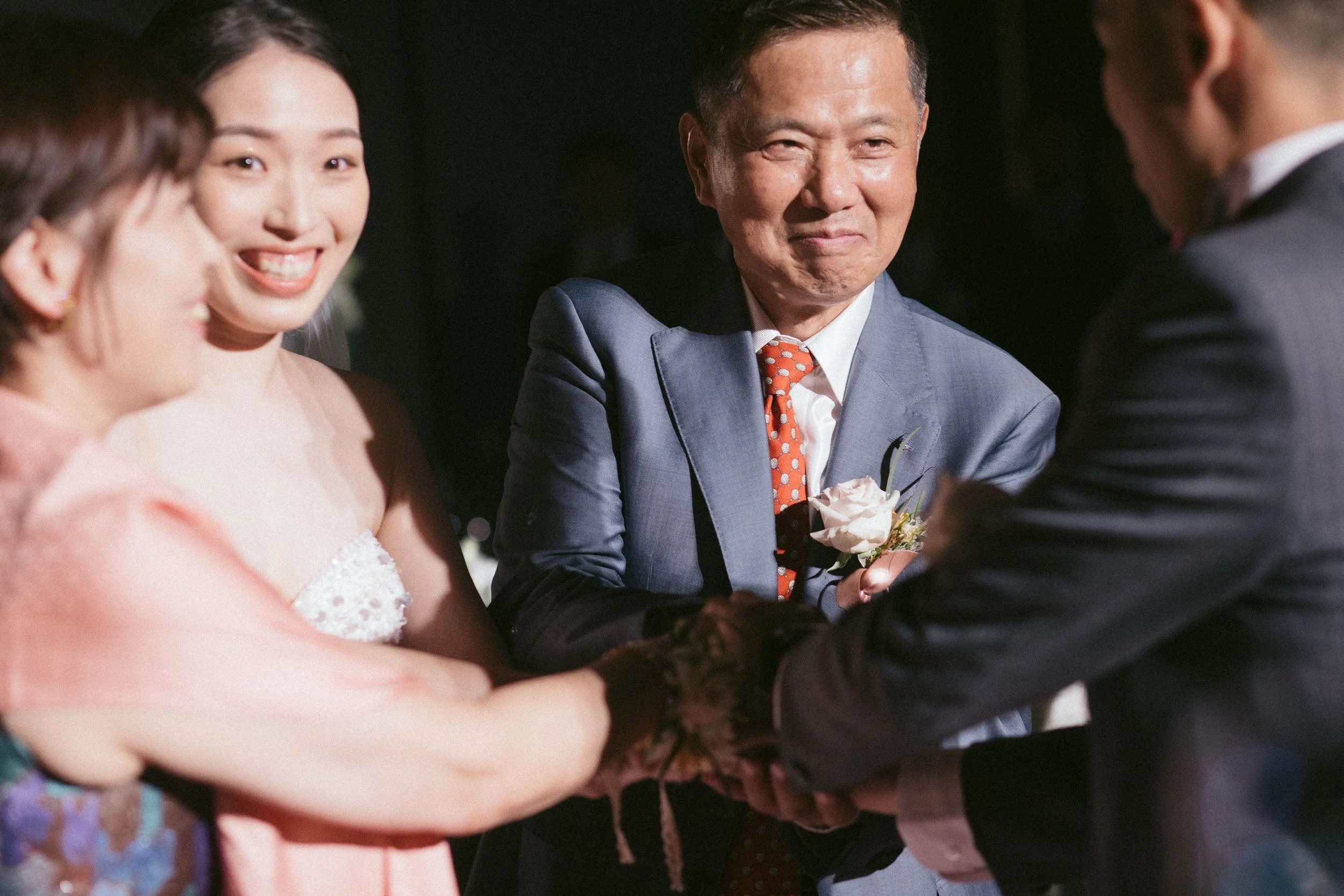 A group of people at a social gathering, including a smiling man in a light blue suit and orange polka dot tie holding a flower. Others are partially visible, engaged in conversation.