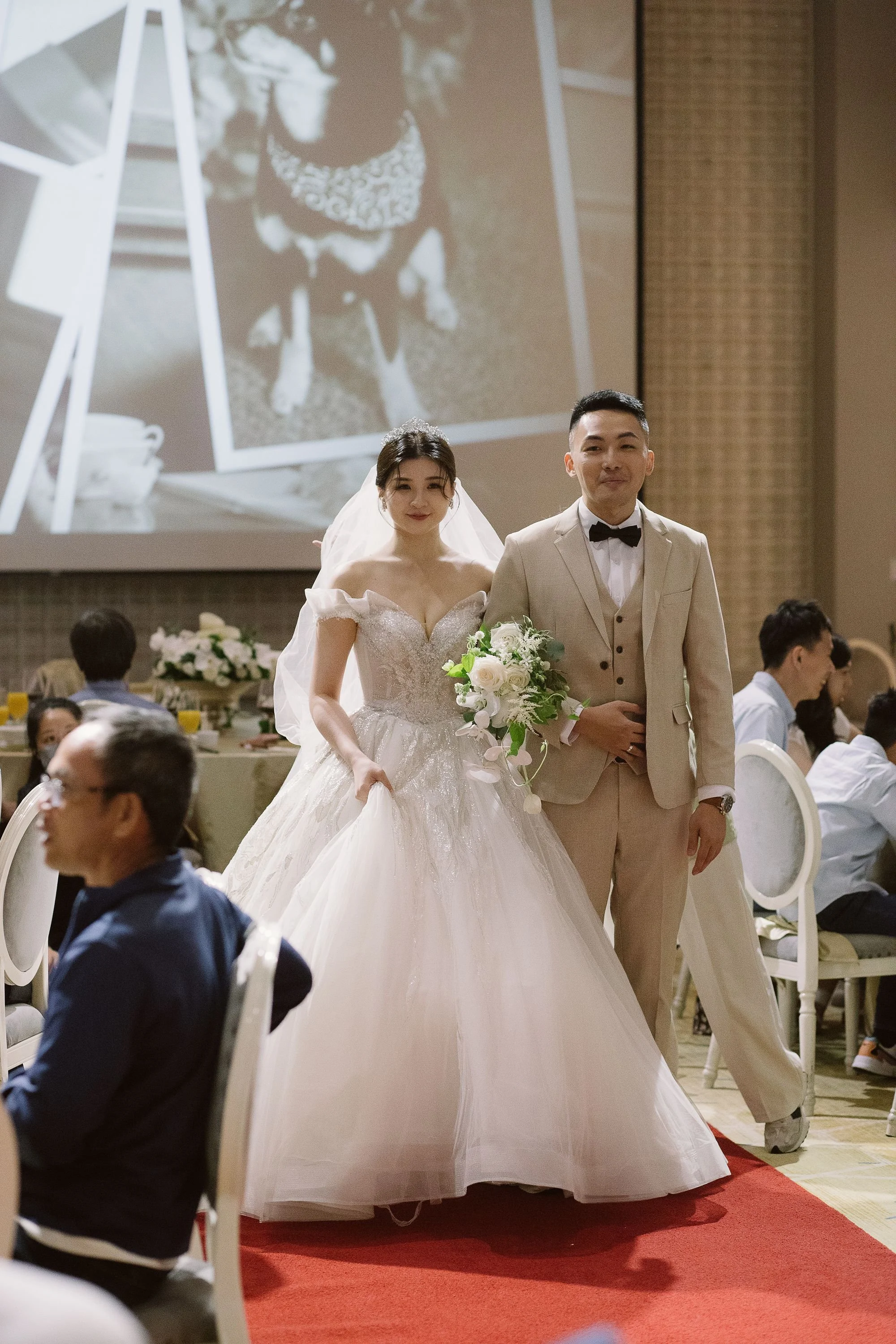 A bride and groom walking down the aisle at their wedding ceremony in a large banquet hall. The bride is wearing a white wedding gown and holding a bouquet of white flowers. The groom is dressed in a light-colored suit with a black bow tie. There are