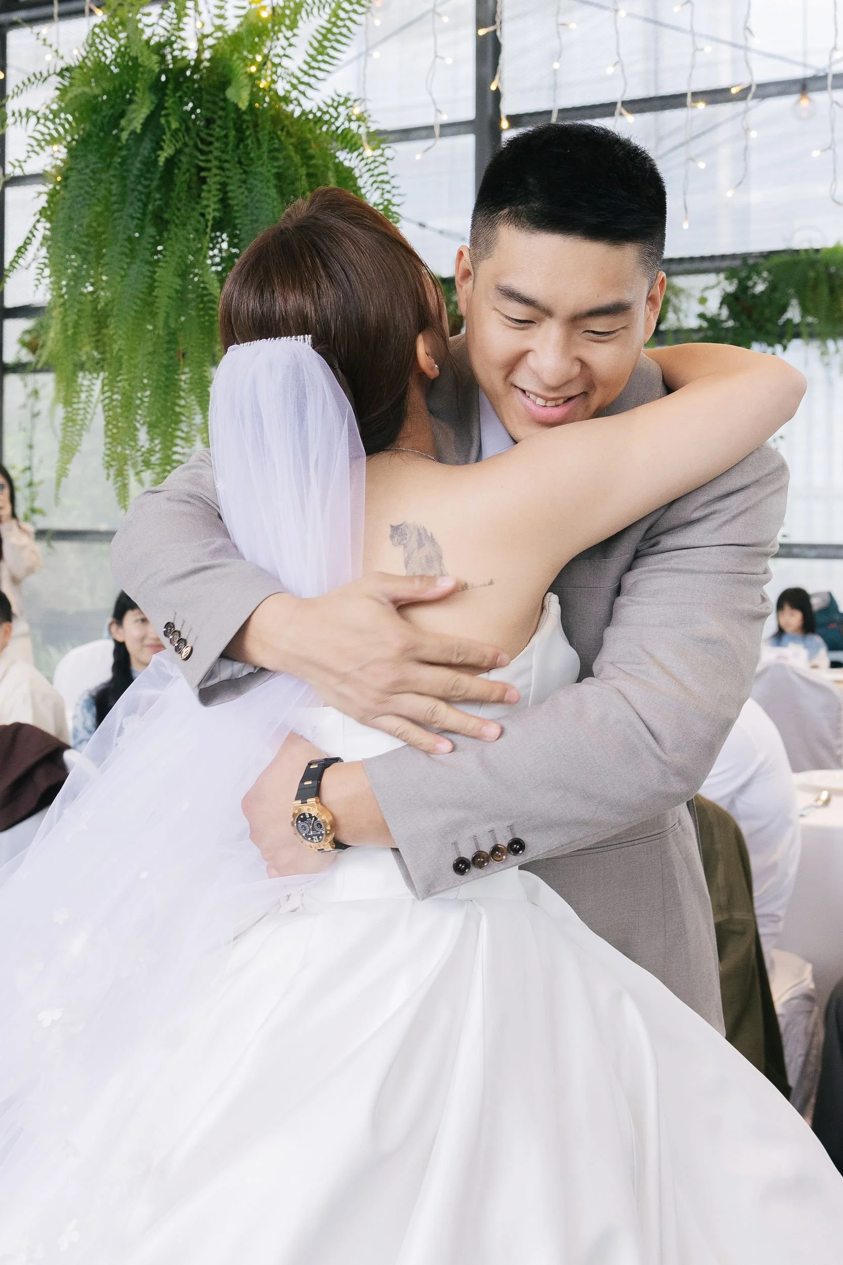 A bride and groom embrace during their wedding reception inside a glass greenhouse venue with hanging fairy lights and green plants. The bride's arm features a tattoo of a horse's head.