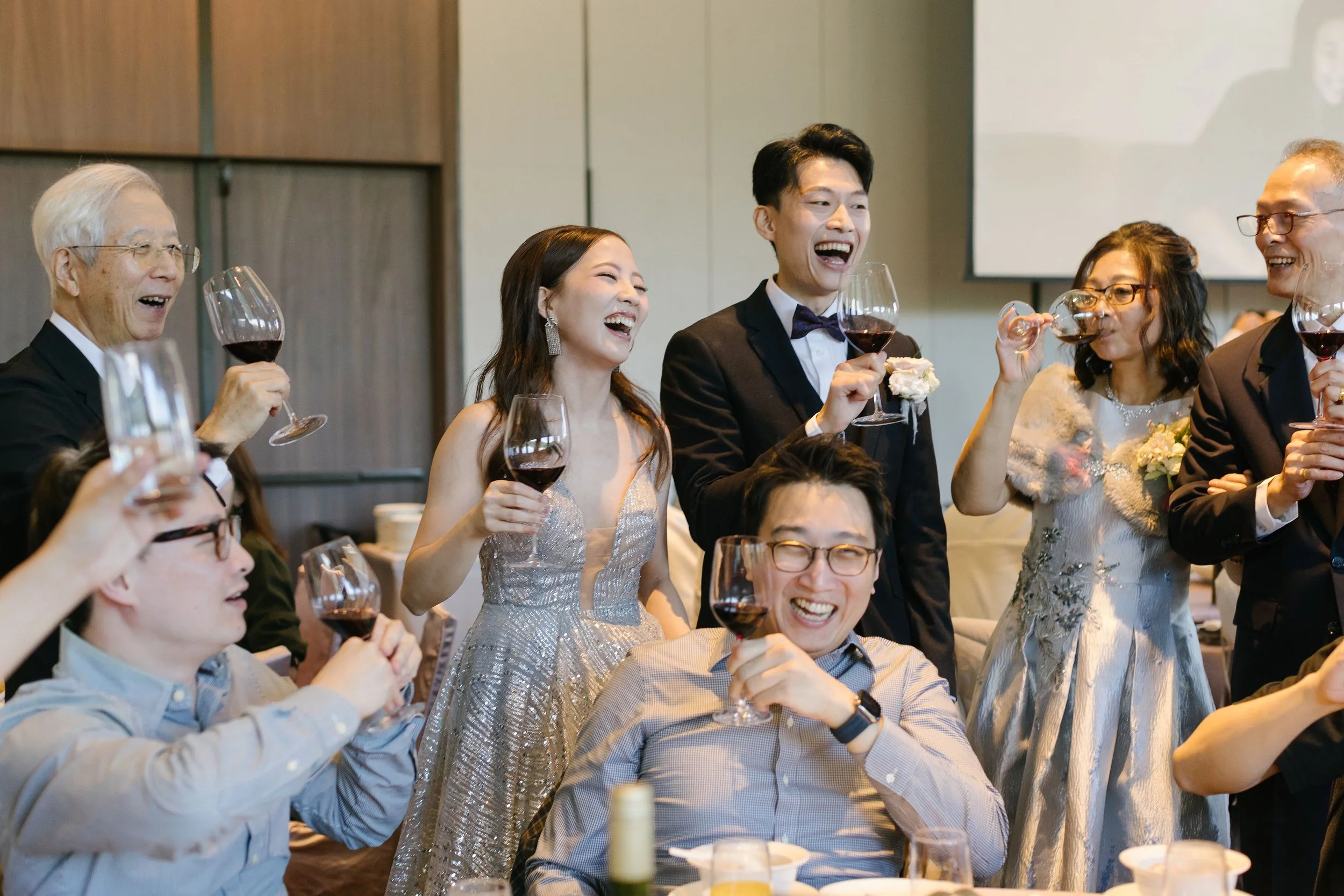 A group of people at a celebration, wearing formal attire, toasting with glasses of red wine, laughing and enjoying themselves.