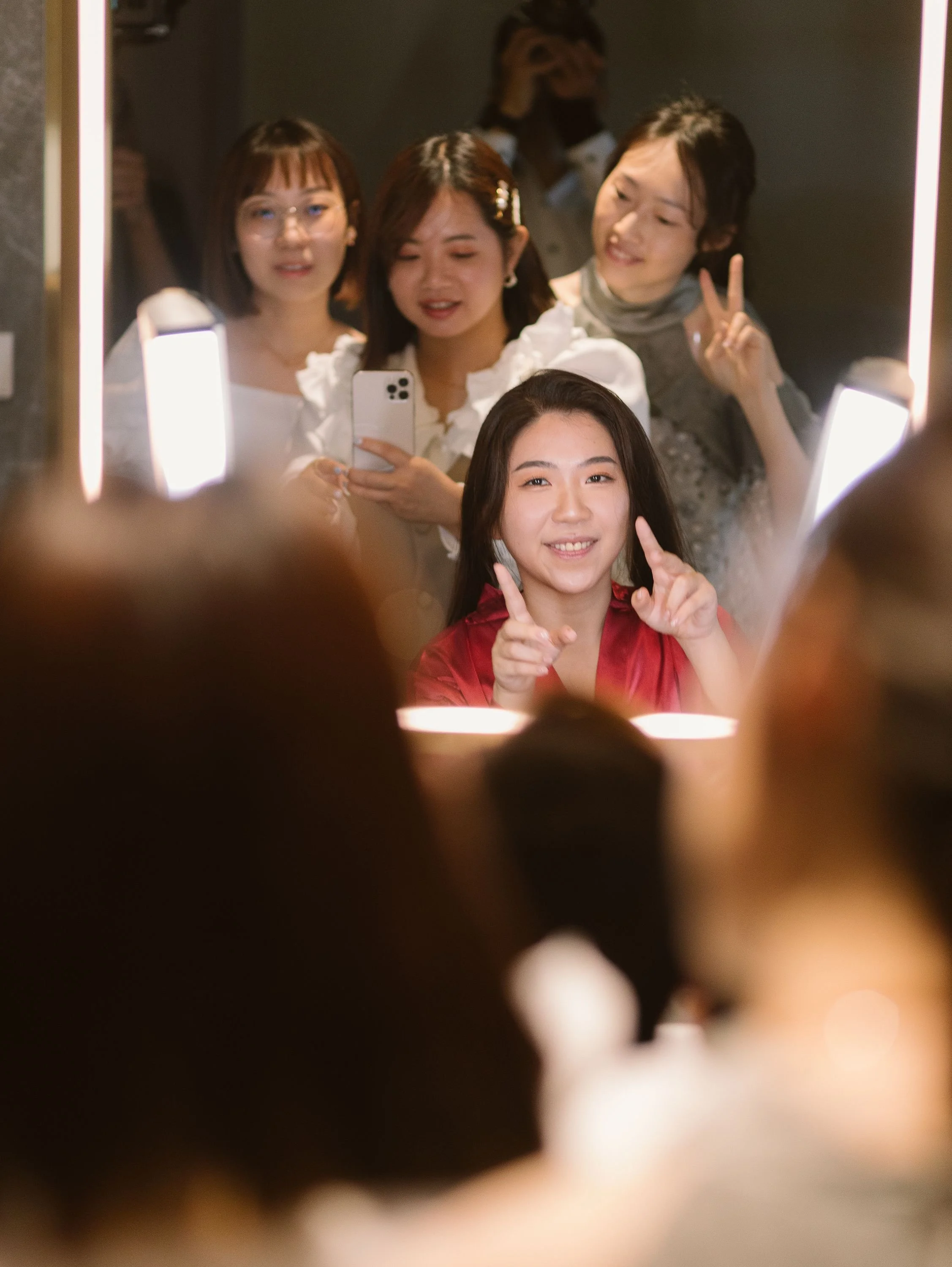 Four women posing for a mirror selfie in a dimly lit room, with one woman holding the phone and everyone smiling or making peace signs.
