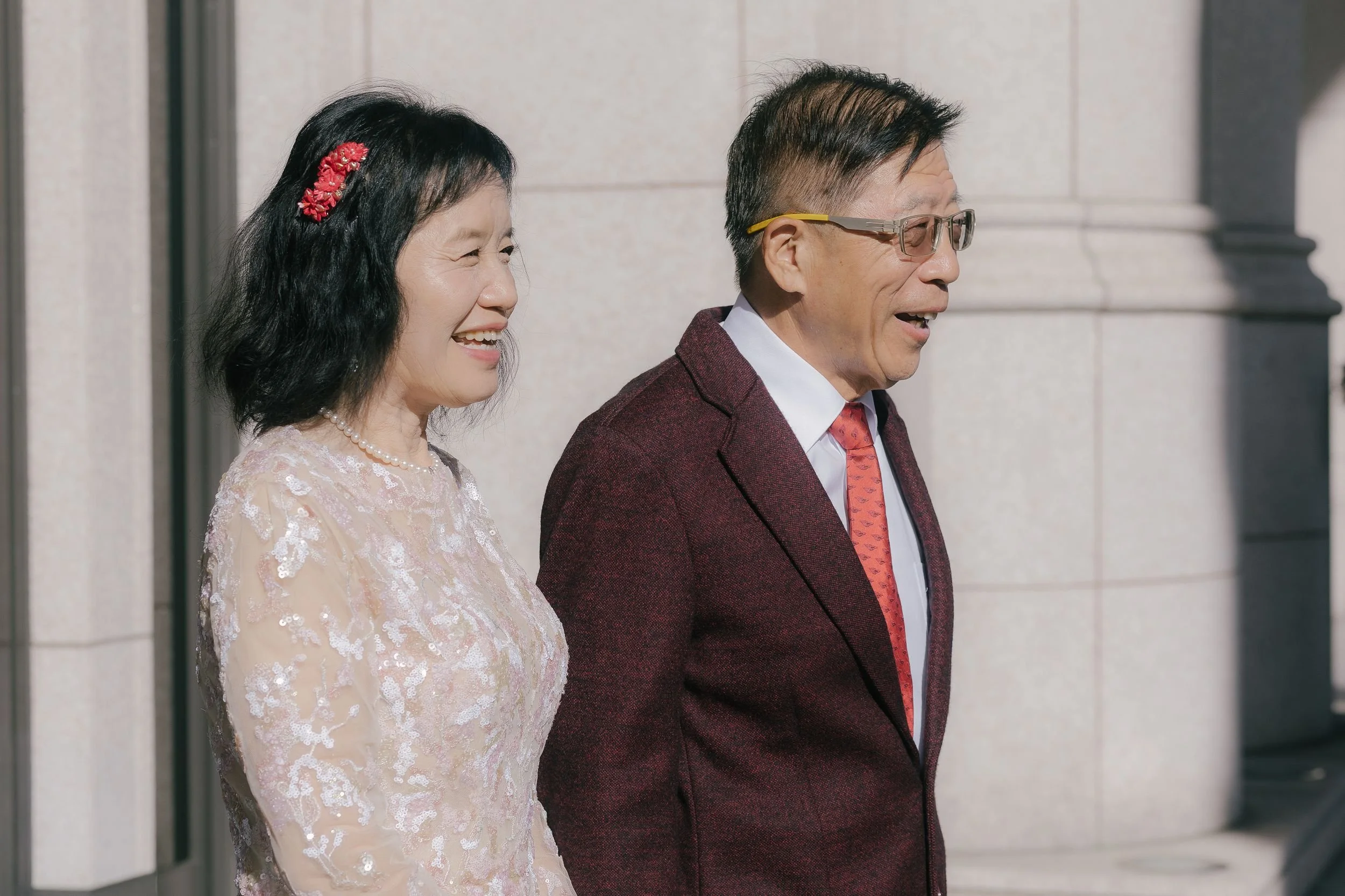 A smiling older Asian woman with black hair decorated with a red flower hair accessory, wearing a light pink lace dress and a pearl necklace, standing next to an older Asian man with short black hair, wearing glasses, a white shirt, a red tie, and a 