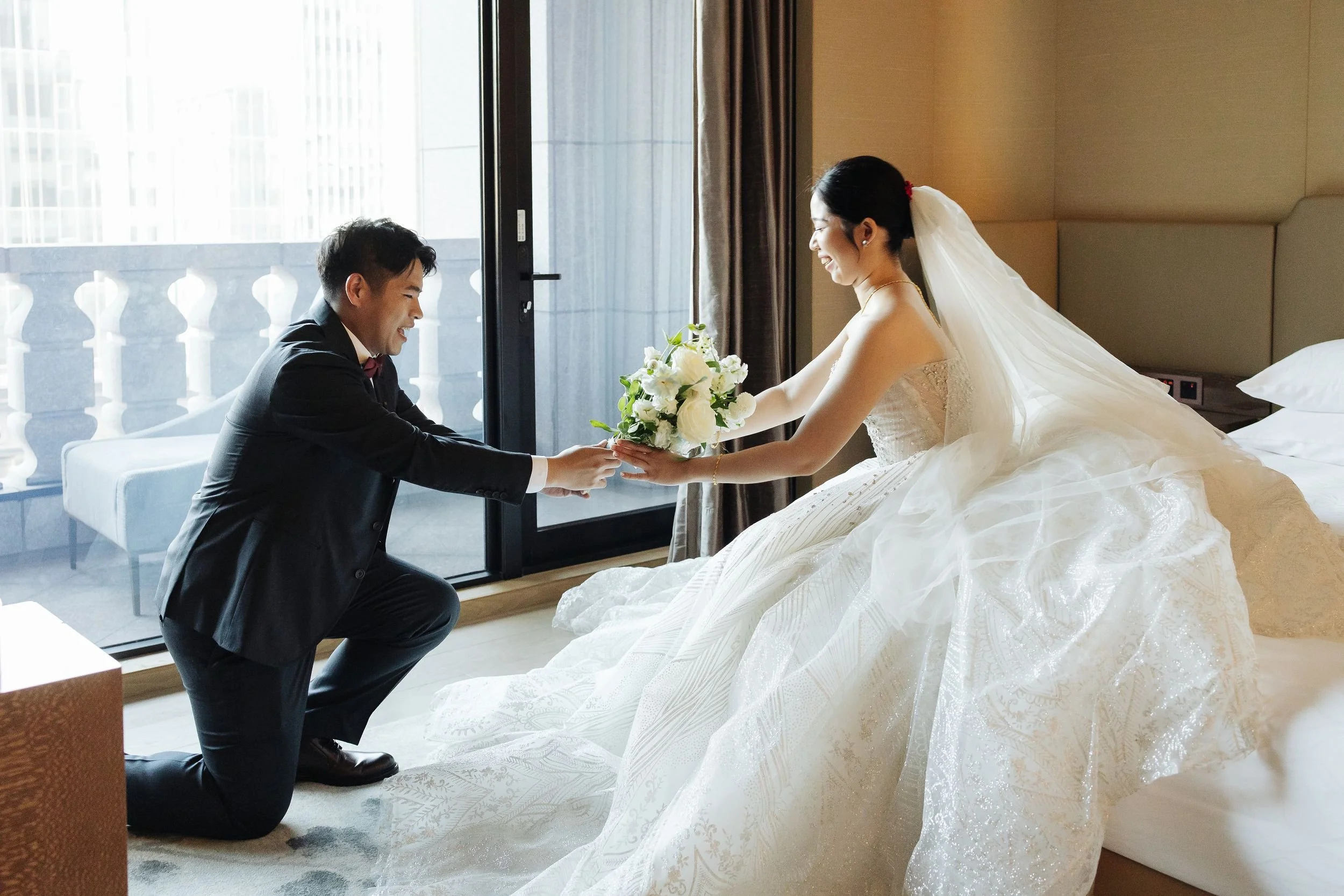 A groom kneeling and holding a bride's hands as she smiles with a bouquet of white flowers, inside a hotel room near a balcony door.
