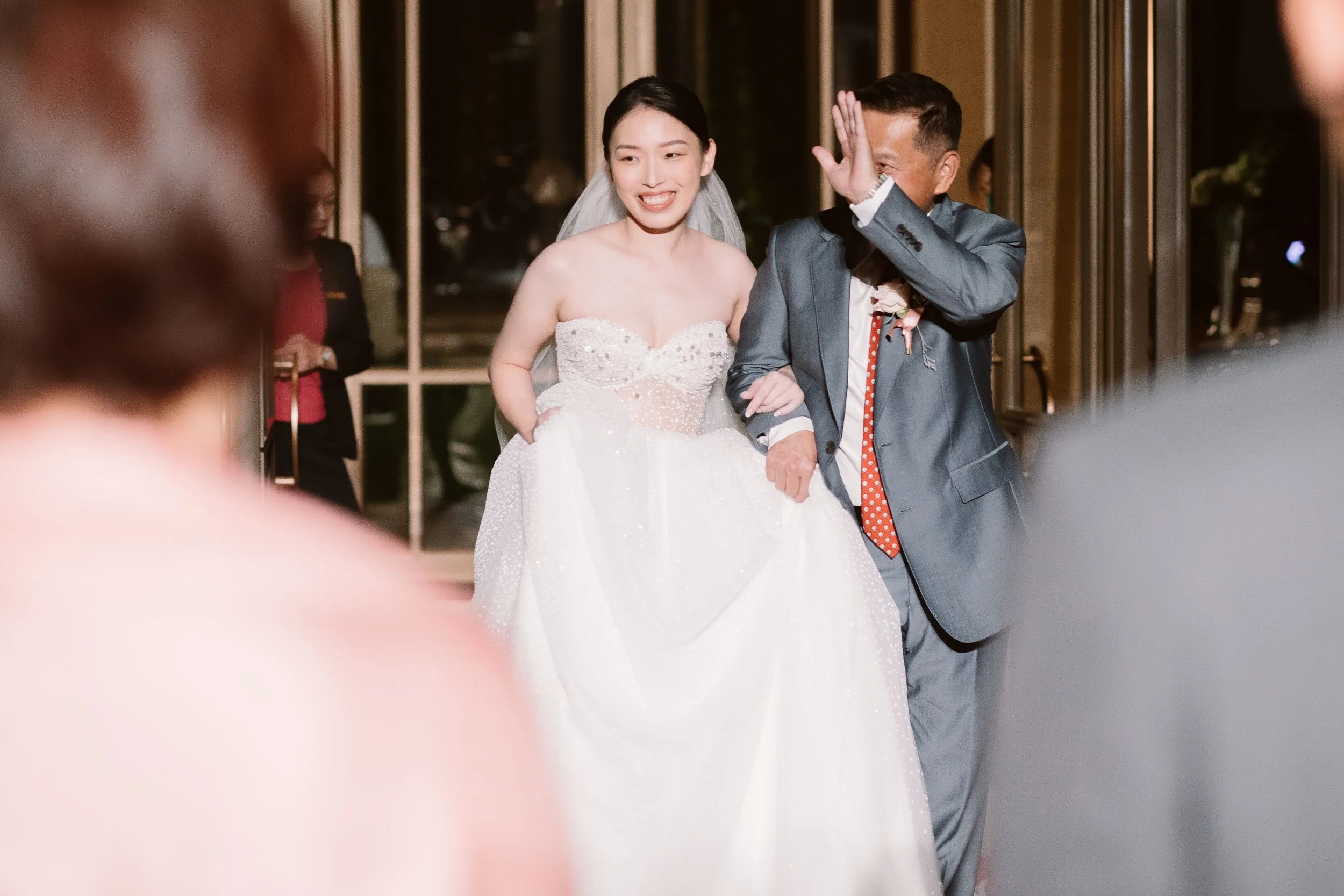 A bride smiling and holding hands with a man, possibly her father, at her wedding. The man covers part of his face with his hand. They are indoors, and other people can be seen in the background.