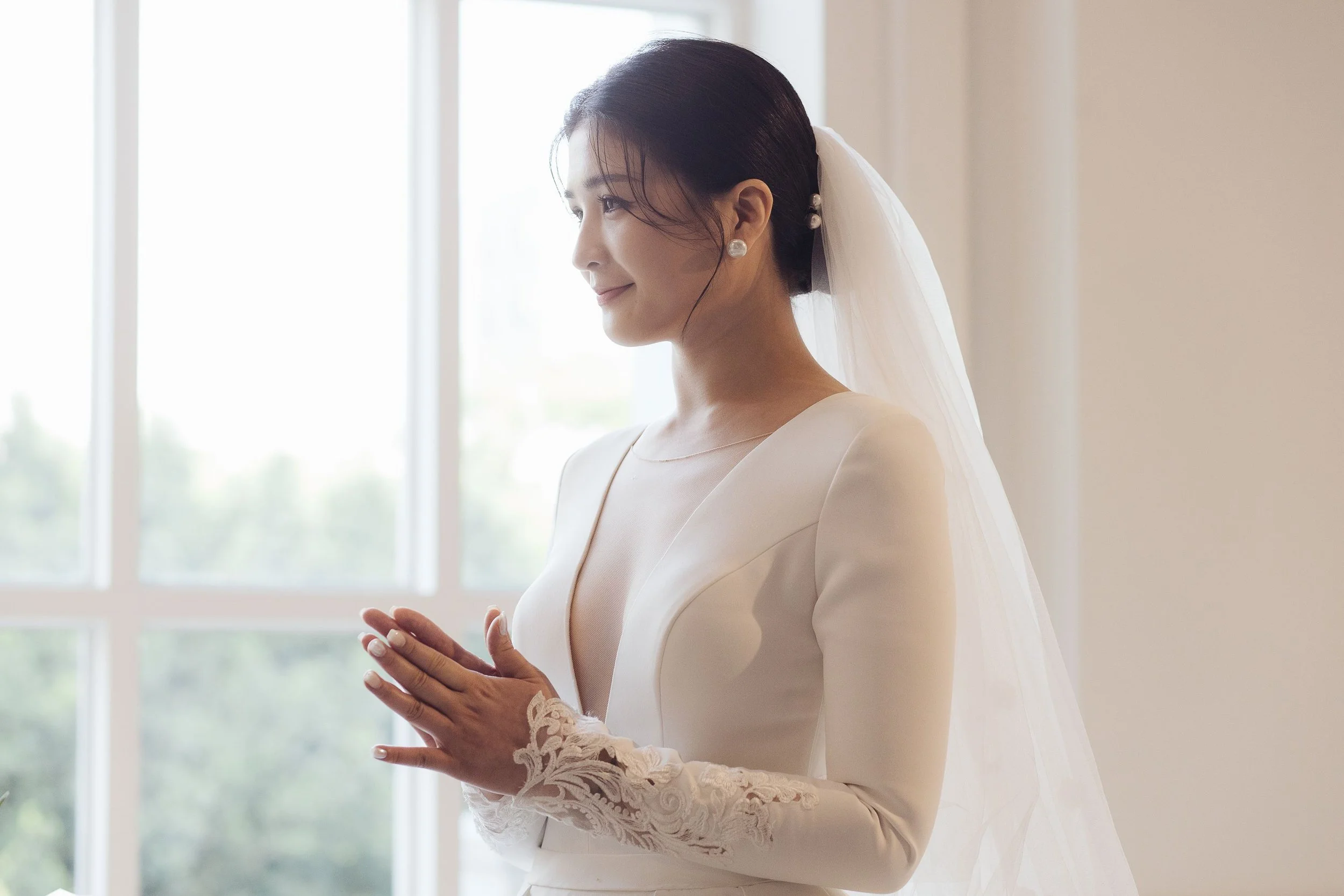 A bride in a white wedding gown with lace sleeves, standing by a large window, with her hands clasped together, looking content and serene.