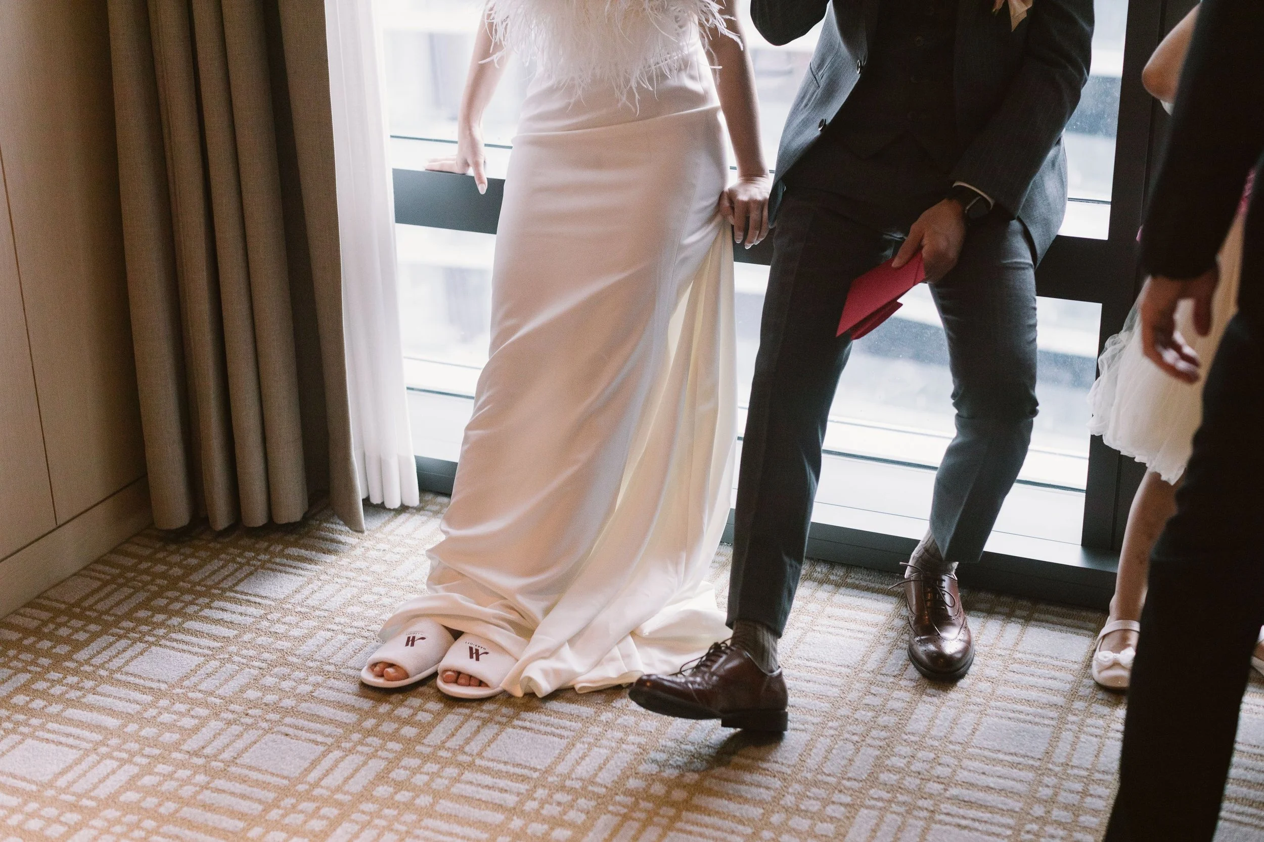 Close-up of people standing indoors near a large window, with one person wearing a white wedding dress and another in a dark suit, holding a red object, possibly a folder or envelope.