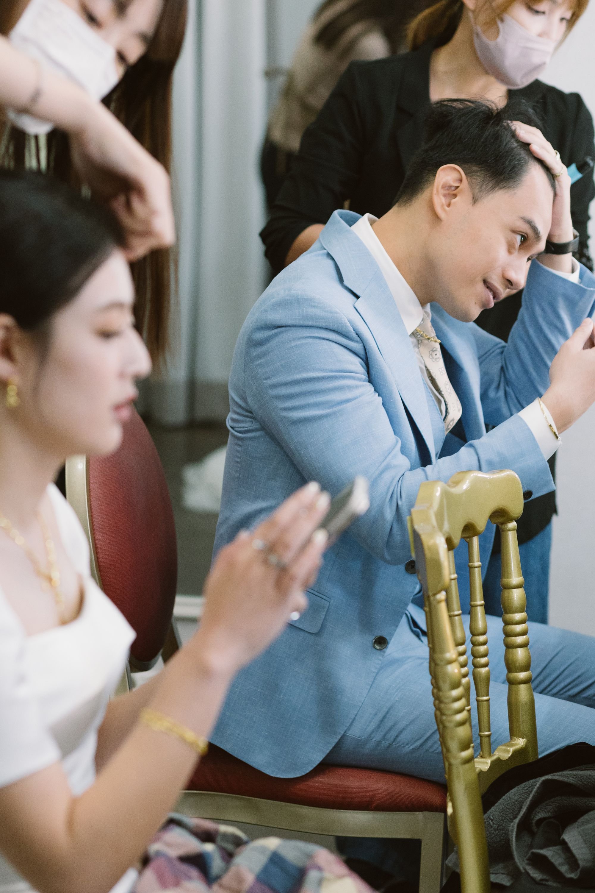 People sitting in a row, wearing face masks, looking at their phones, with gold-colored chairs, in an indoor setting.