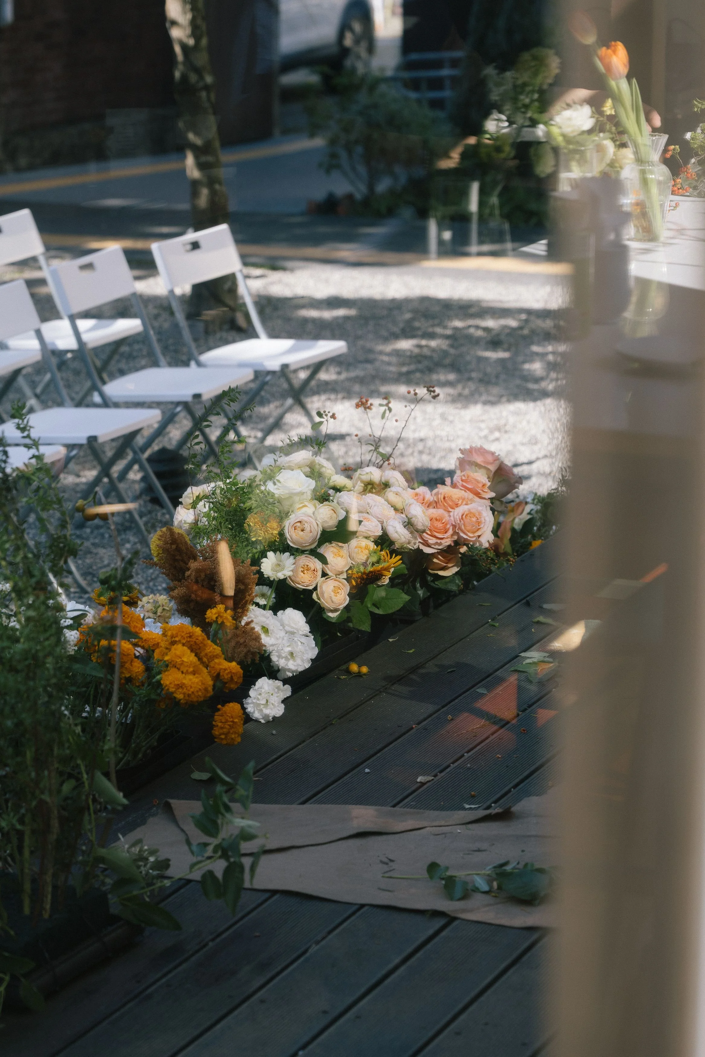 View of a display of various flowers, including roses and marigolds, arranged on a black wooden surface. Outside, in the background, there are white chairs and a gravel path, with a partially visible person and parked car reflecting in a glass window