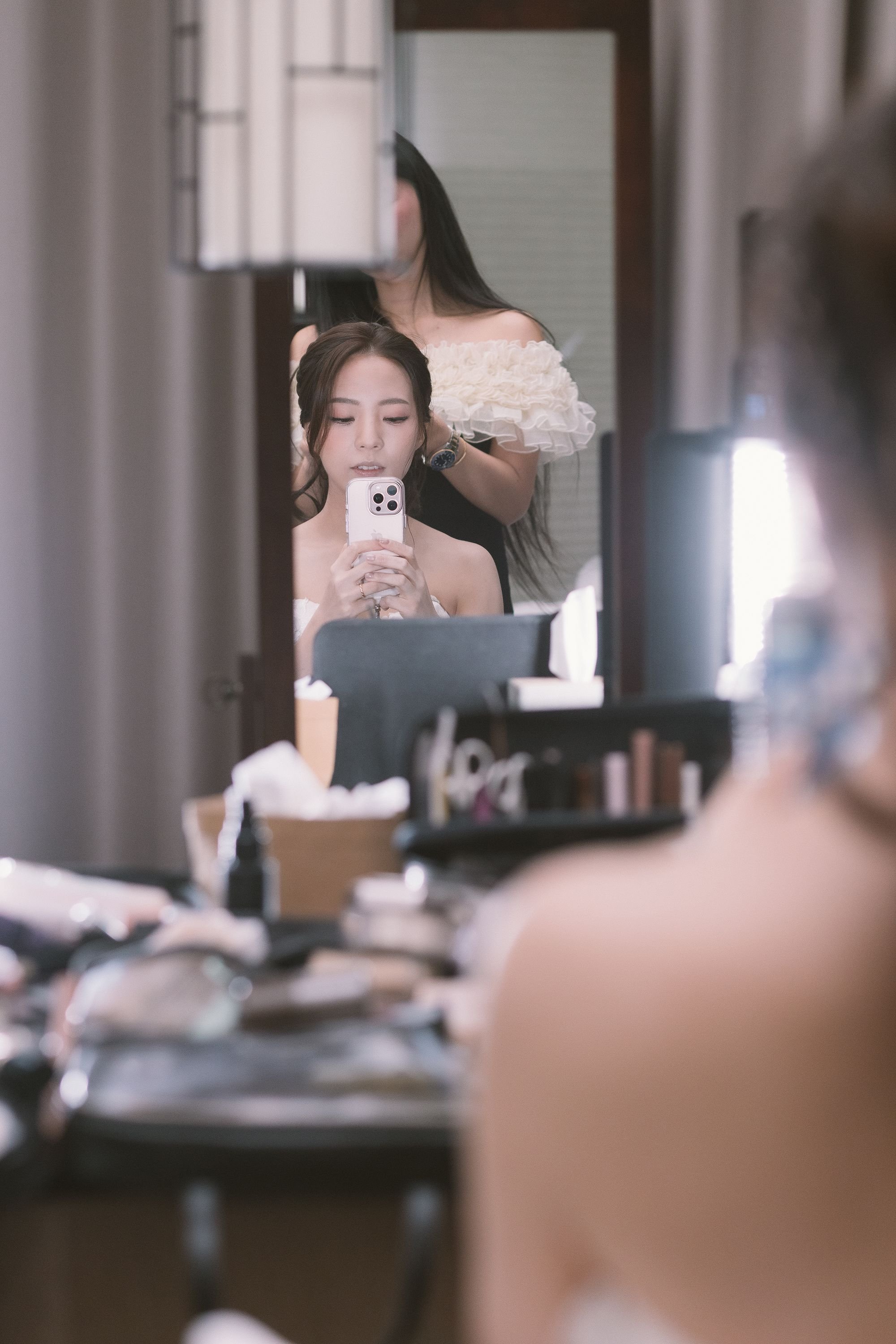 A woman is taking a mirror selfie while another woman is doing her hair in the background. The scene appears to be a dressing or makeup area with various cosmetic products and a makeup mirror visible.