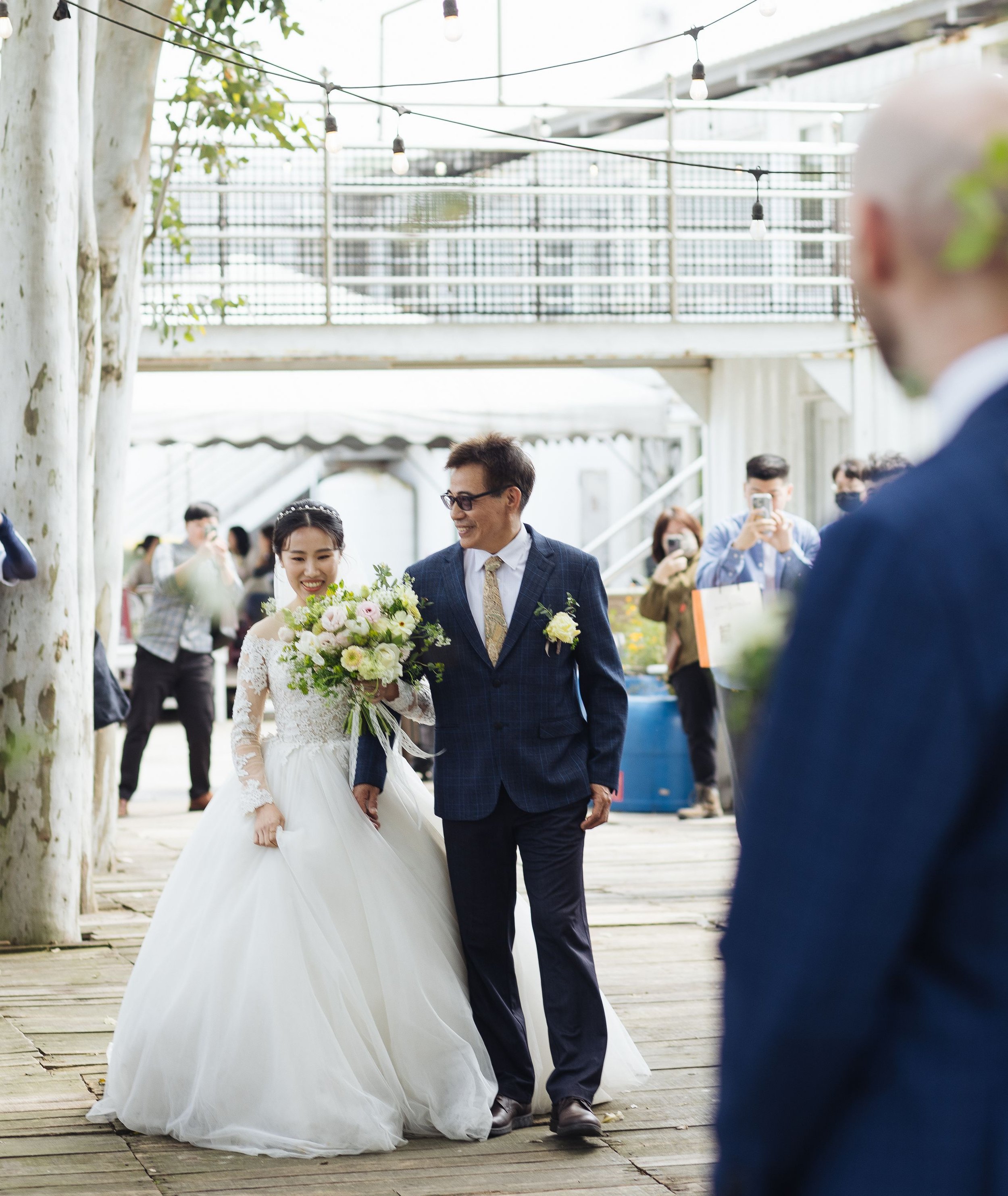 A bride in a white wedding dress and a groom in a suit walk together, surrounded by guests at an outdoor wedding celebration. The bride holds a bouquet of flowers and the scene is lively with some guests taking photos or observing.