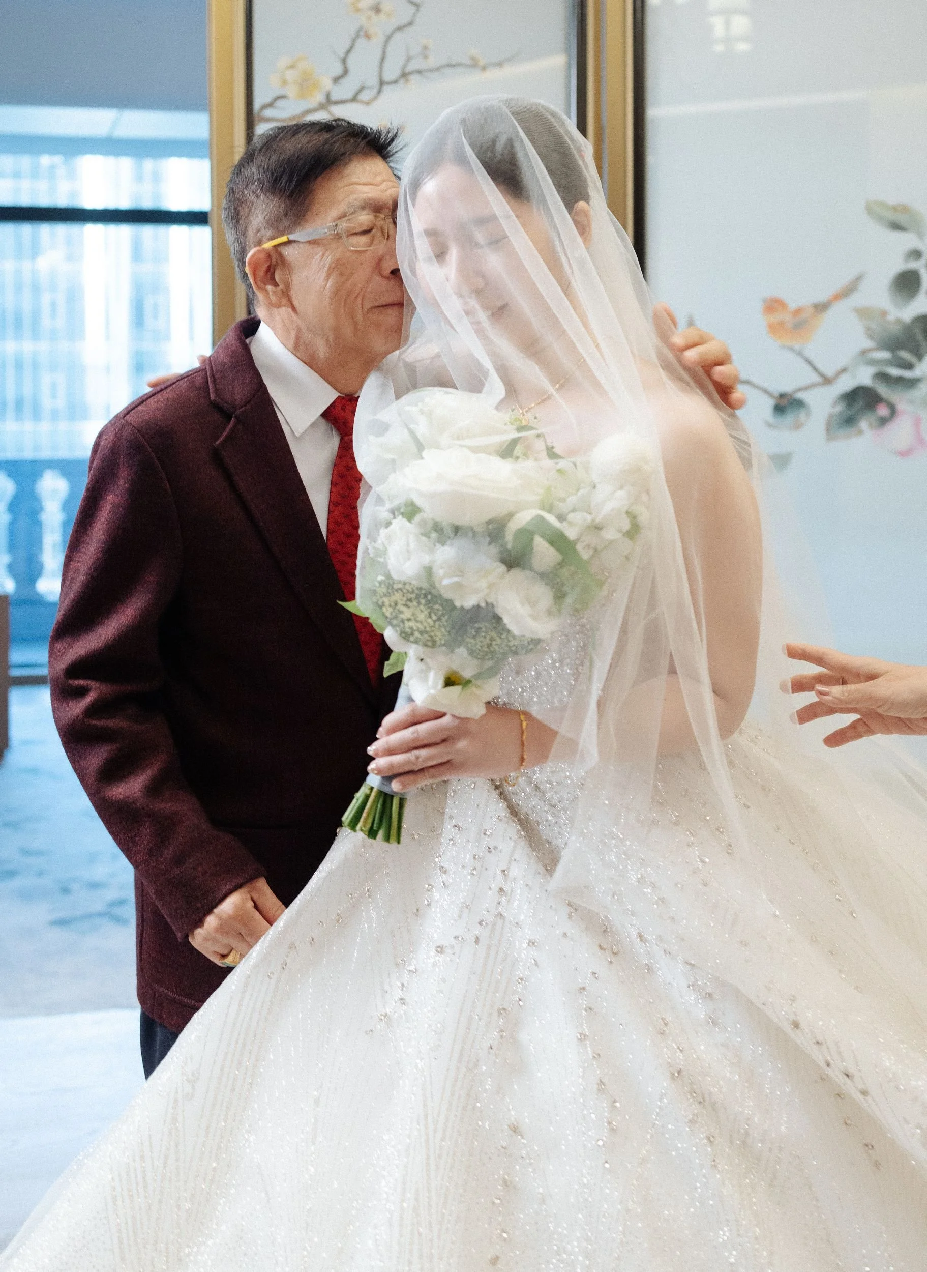 A groom dressed in a burgundy suit embraces a bride in a white wedding gown holding a bouquet of white flowers, as they share a moment with their faces close together in an indoor setting with decorative artwork in the background.