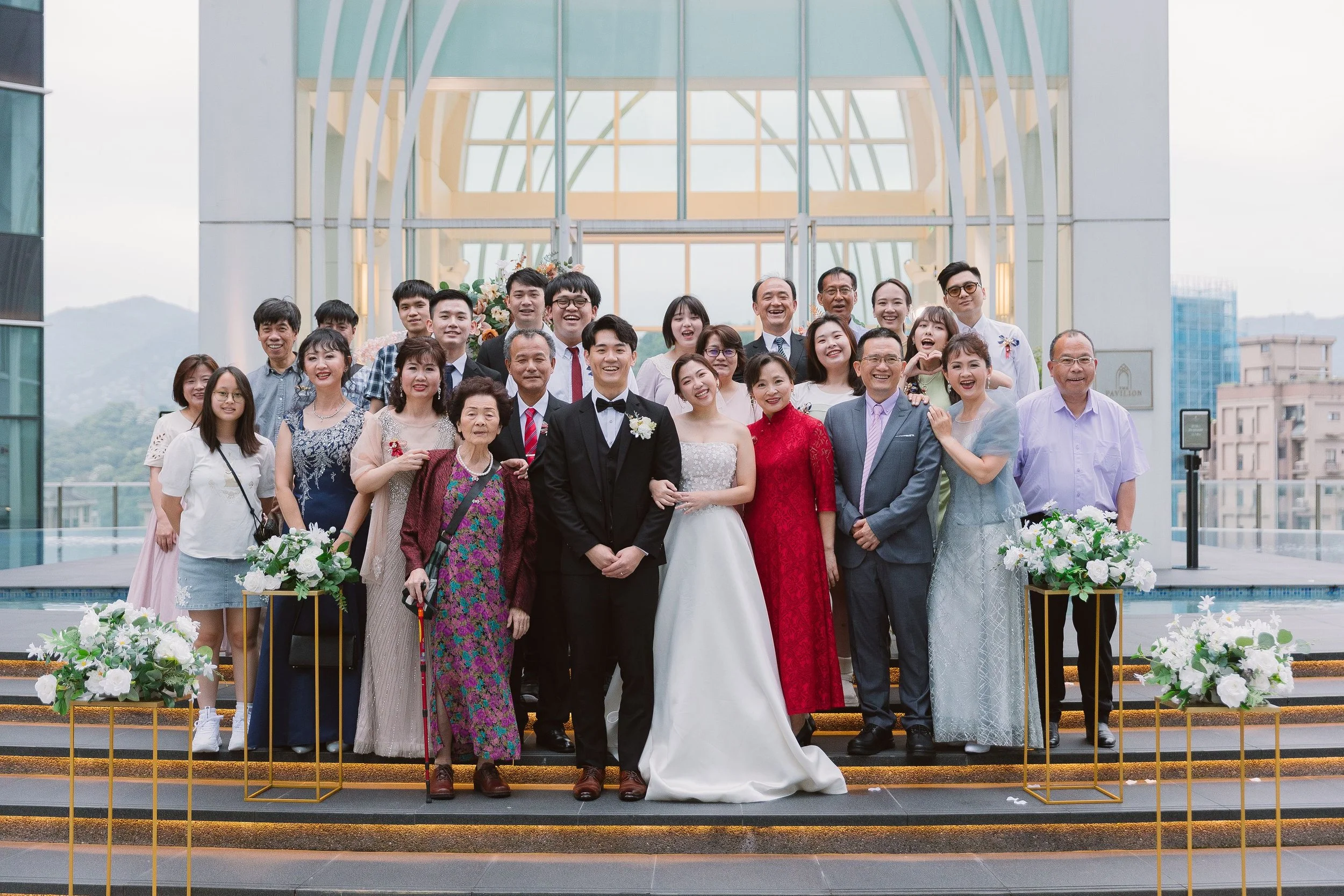 A large group of people gathered for a wedding celebration outdoors, with the bride and groom in the front center, surrounded by family and friends smiling and posing for a photo.
