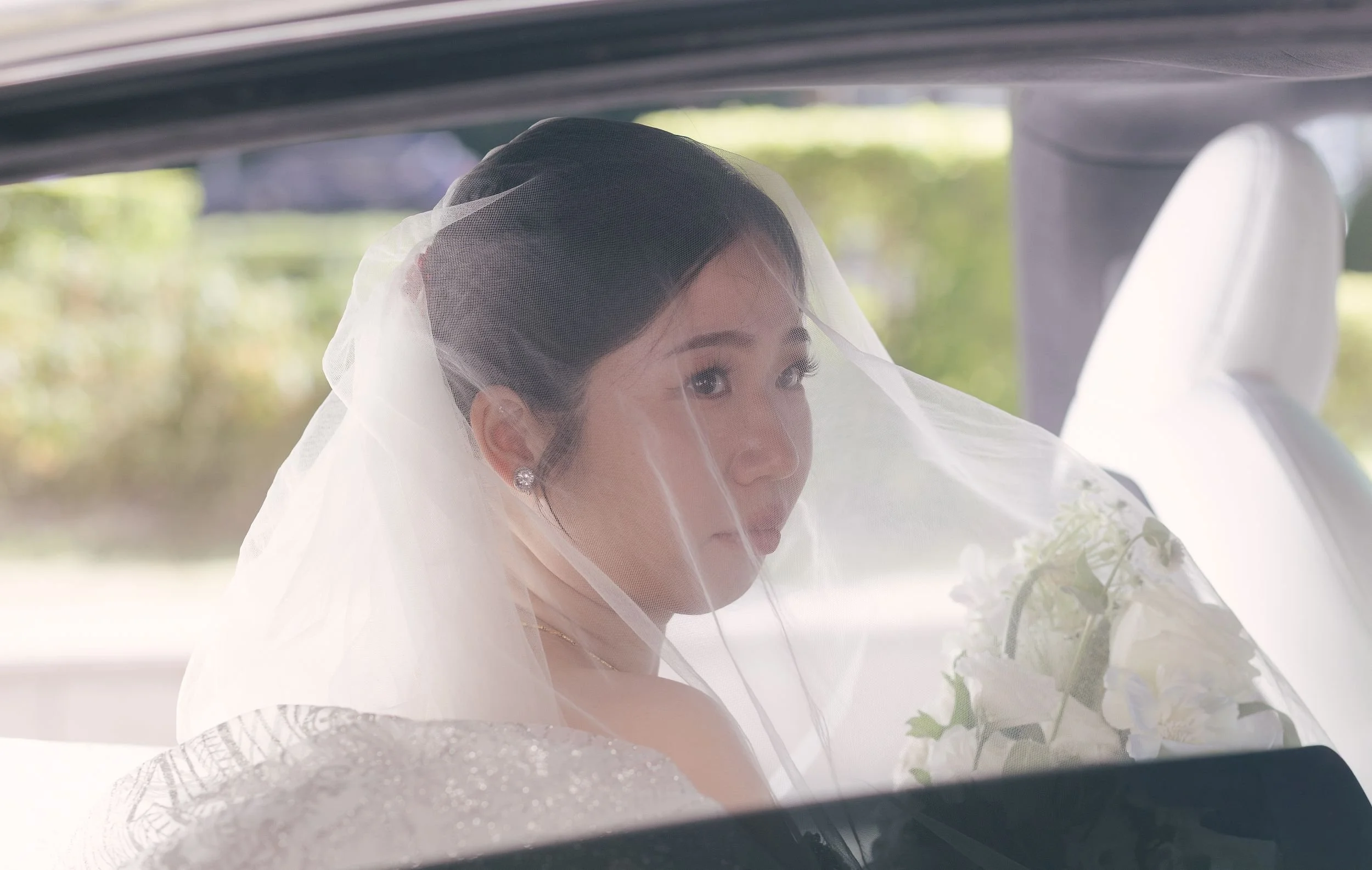 A bride with dark hair styled in an updo, wearing a white wedding dress and veil, sitting in a vehicle with a blurred outdoor background.