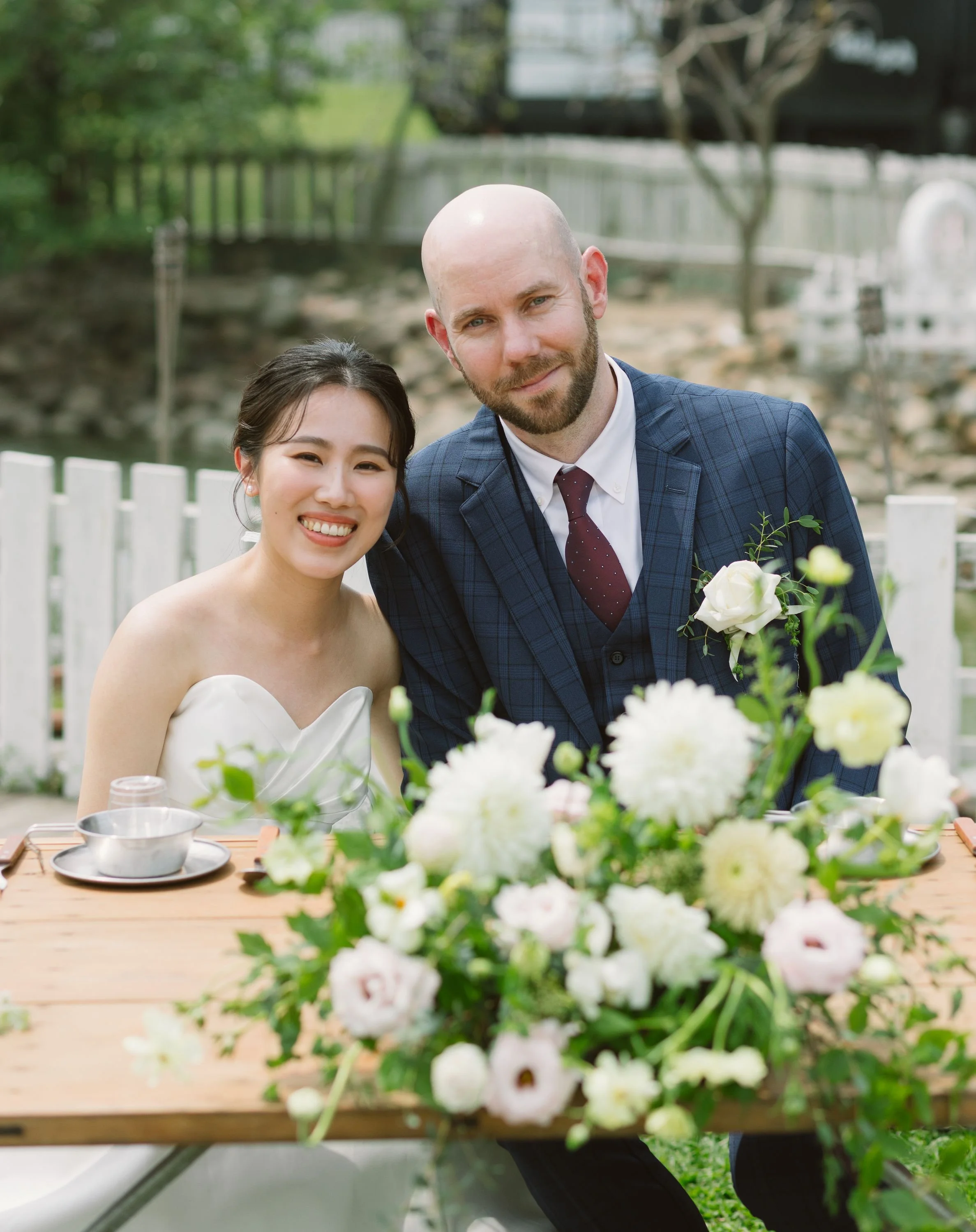 A smiling Asian woman and a man with a beard, both dressed formally, at a wedding reception outdoors with a garden setting, a table with floral arrangements, white fence, and some trees in the background.