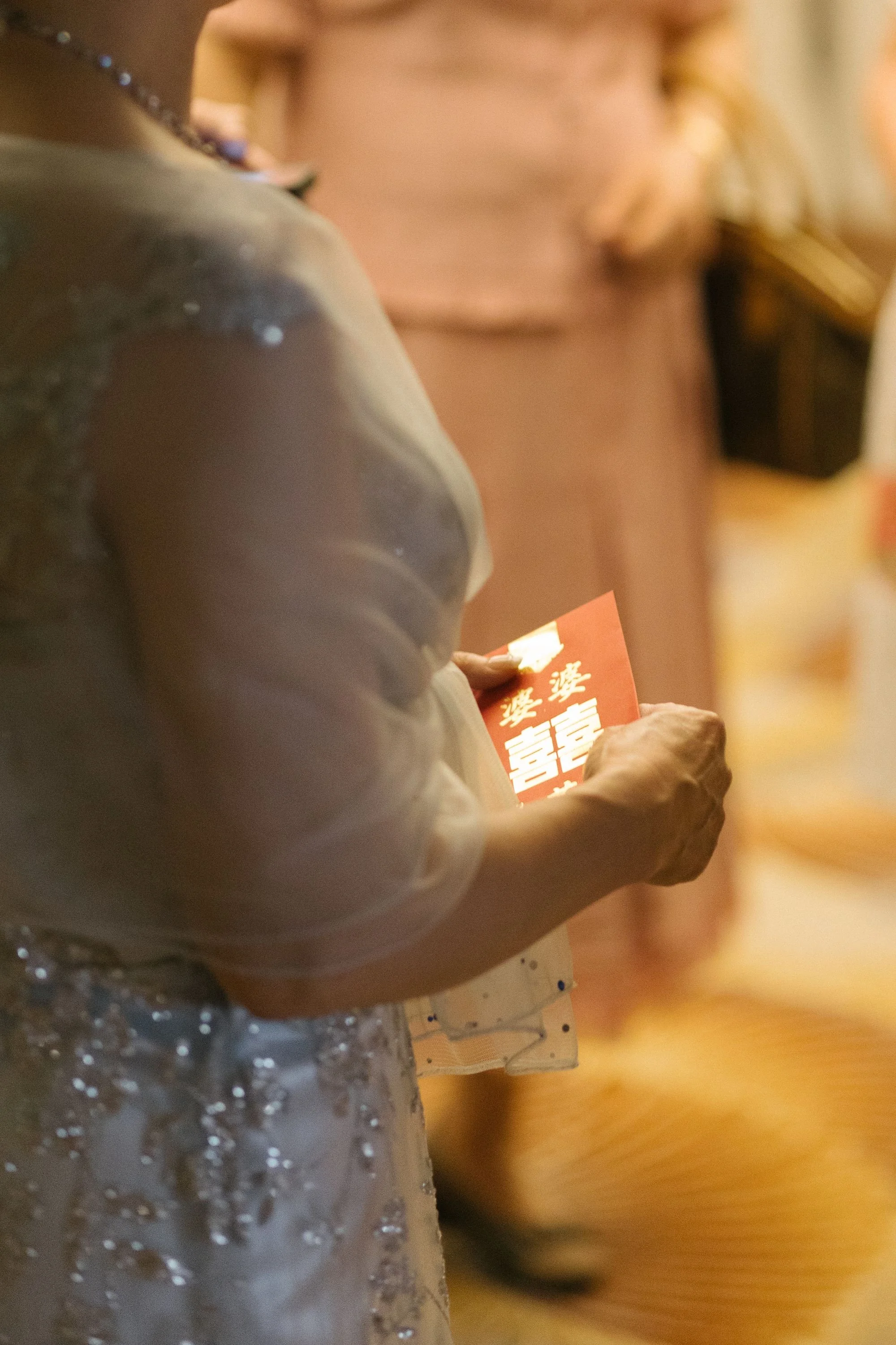 Close-up of an elderly woman holding red envelope with Chinese characters, dressed in a light-colored, sparkly dress.