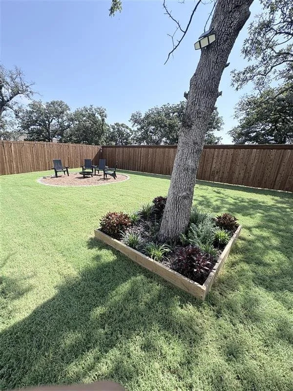 A backyard with a new wooden fence, a large tree surrounded by a planter bed with shrubs and plants, and a circular patio area with three black chairs and a table.