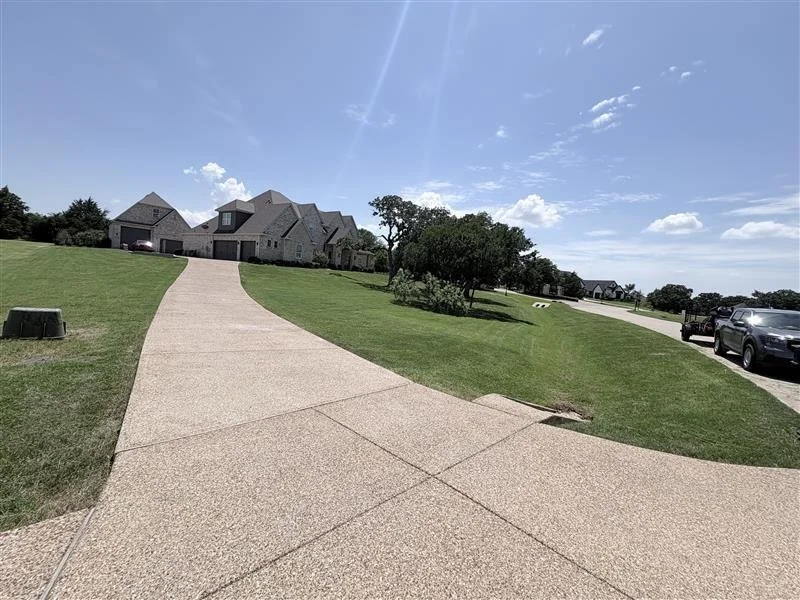 A wide view of a sunny residential neighborhood with a curved sidewalk leading up to a large house, green lawns, trees, parked cars, and a blue sky with some clouds.