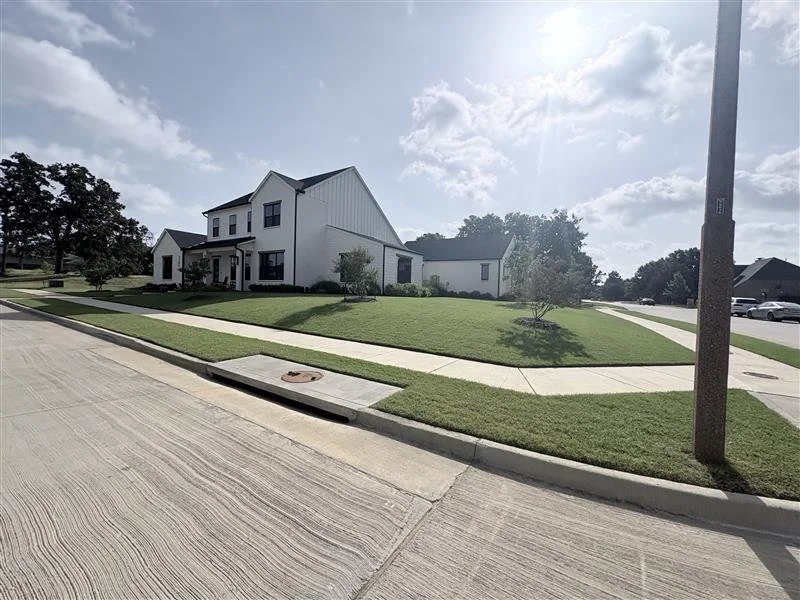 A suburban neighborhood with a modern white house, a neatly mowed lawn, and a sidewalk. The sky is partly cloudy with sunlight streaming through.