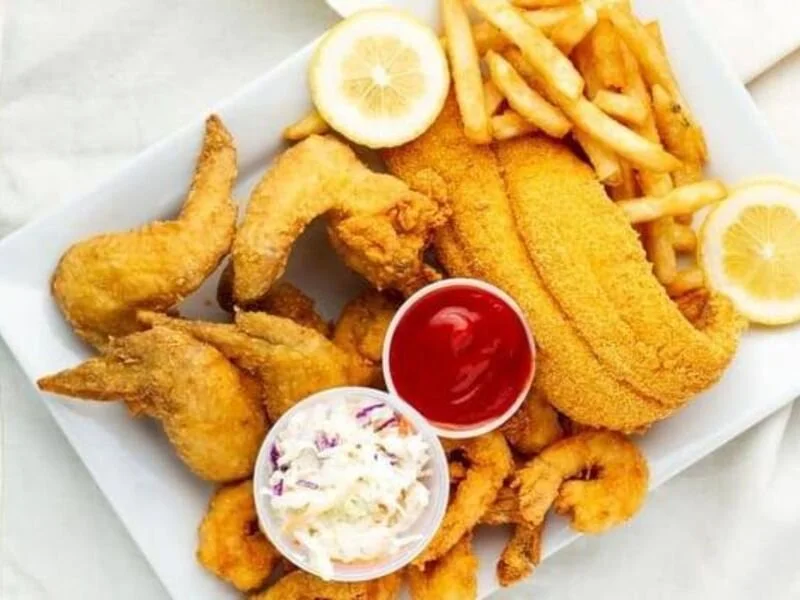 Basket of fried chicken wings, fish fillet, French fries, lemon wedges, and dipping sauces, one of which appears to be ketchup and the other coleslaw.