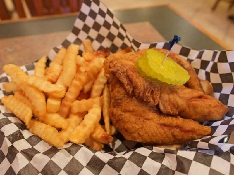 Fried chicken wing with a pickle slice and crinkle-cut French fries served on checkered paper in a basket.