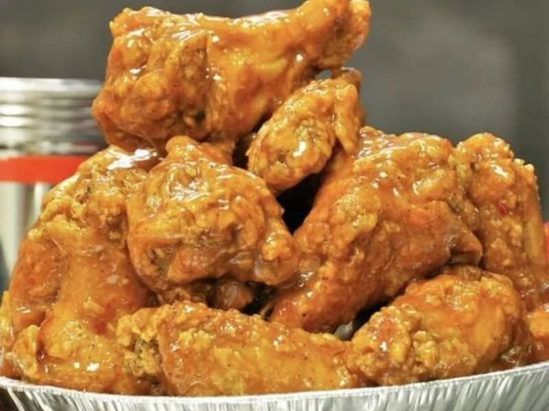 A close-up of a metal tray filled with crispy, golden-brown fried chicken wings covered in sauce.