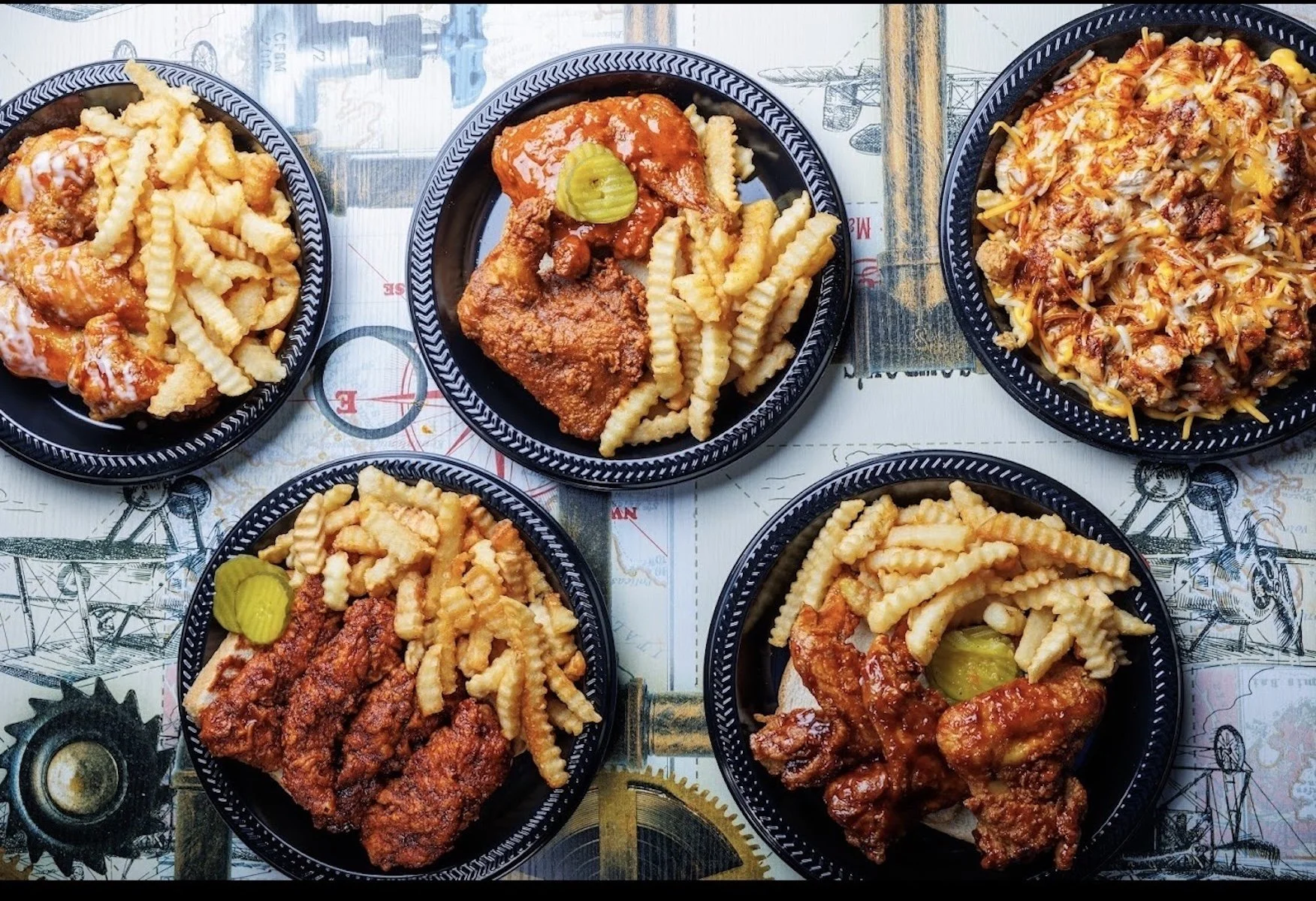Five black oval plates with fried chicken, curly fries, and pickles on a table with an industrial-themed map design.