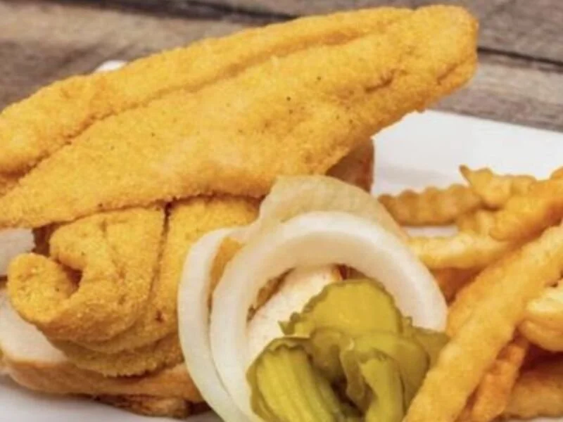 Fried fish fillet sandwich with pickles, onions, and French fries on a plate.