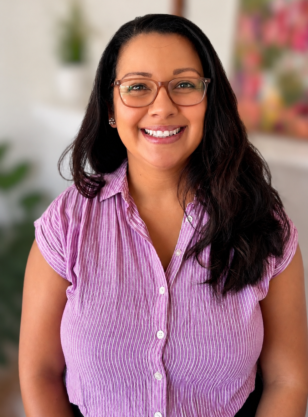 A woman with long dark hair, wearing glasses and a pink striped shirt, smiling at the camera with a blurred colorful background.
