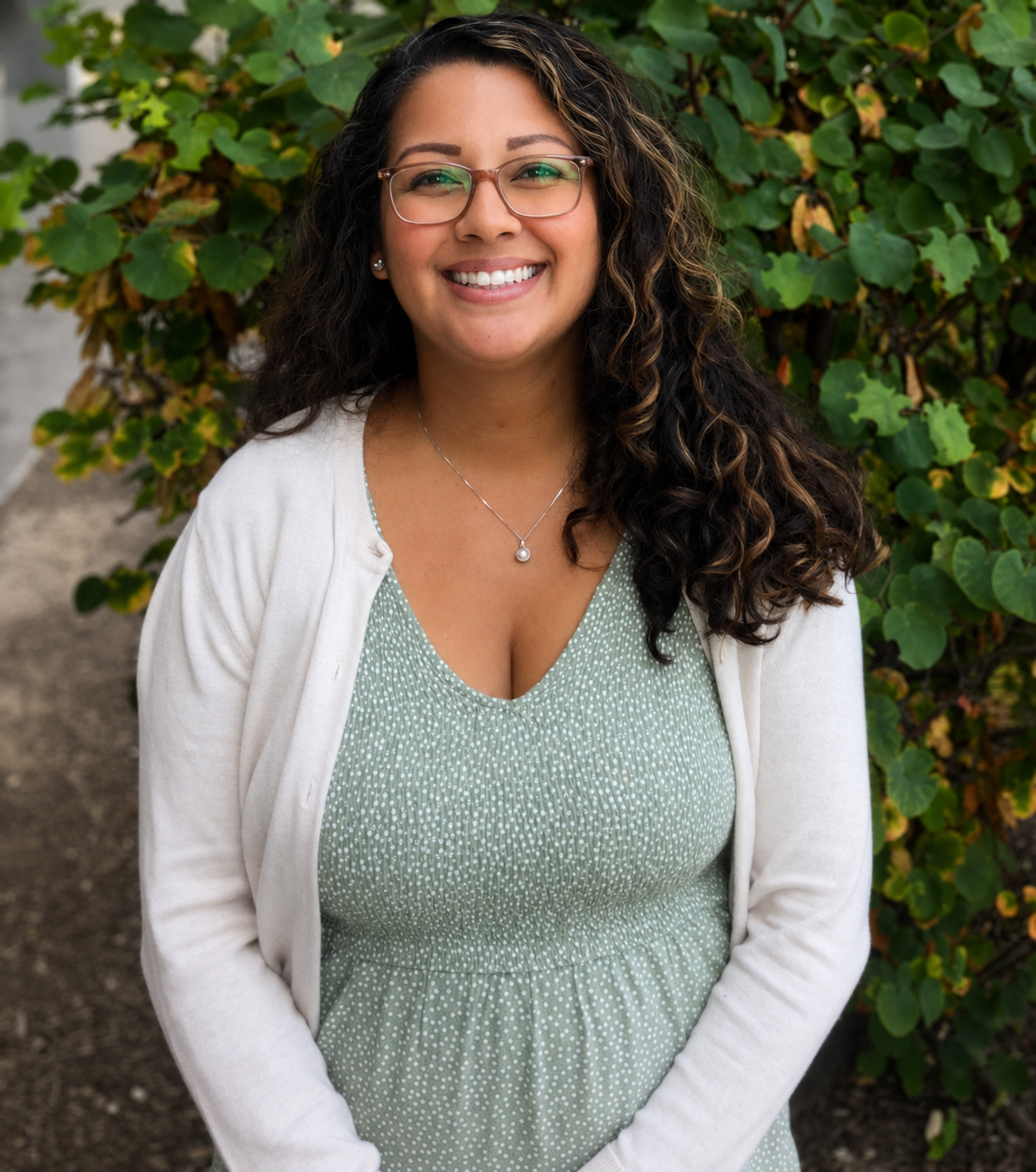 female therapist standing with hands clasped at waist. therapist has dark curly hair with glasses and green dress with beige cardigan