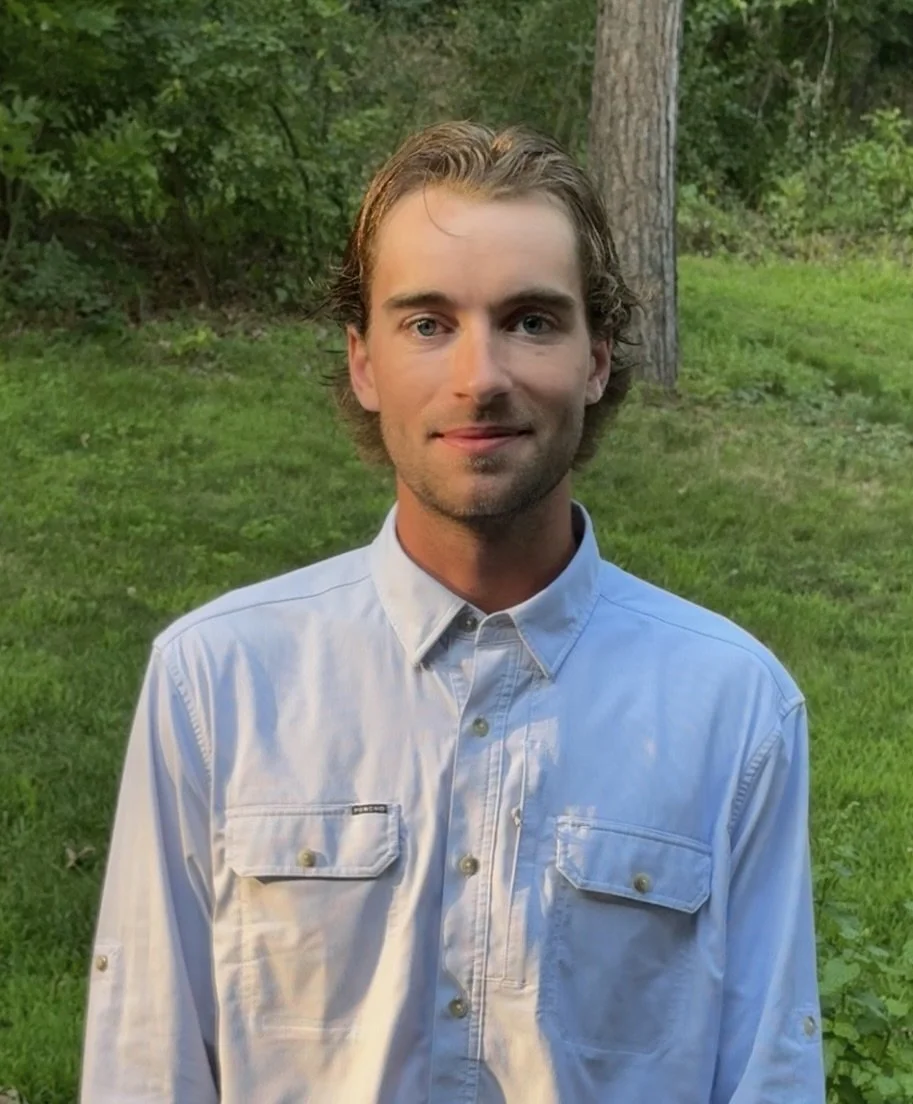 A young man with wavy brown hair, light skin, and blue eyes smiling outdoors with trees and grass in the background, wearing a light blue button-up shirt.