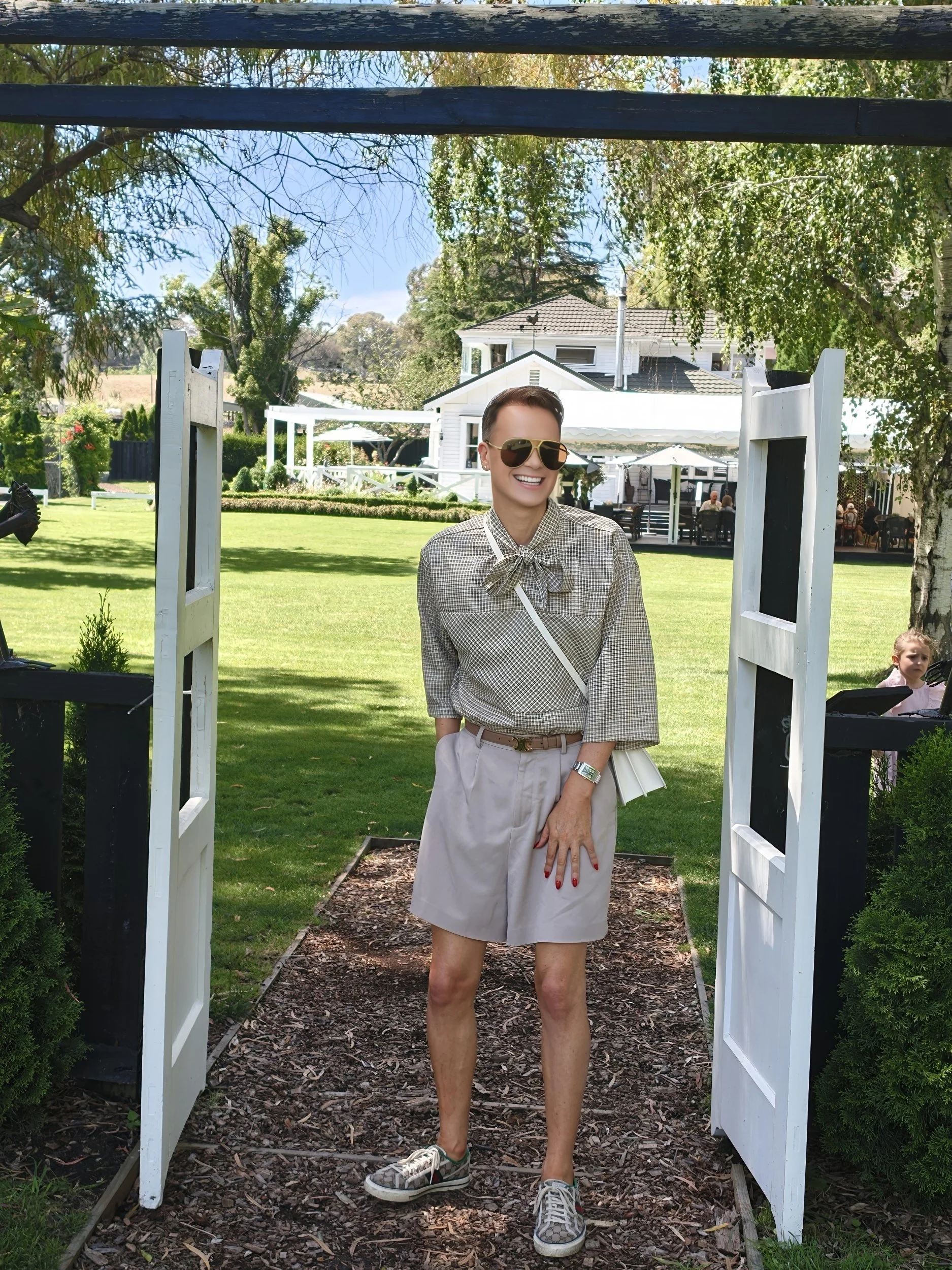 A woman standing outside on a garden pathway, wearing sunglasses, a checkered blouse with a bow, beige shorts, and sneakers. She is smiling and standing in a wooden archway with an open white gate. The background shows a lush lawn, trees, a white house with a gray roof, and outdoor seating areas with umbrellas.