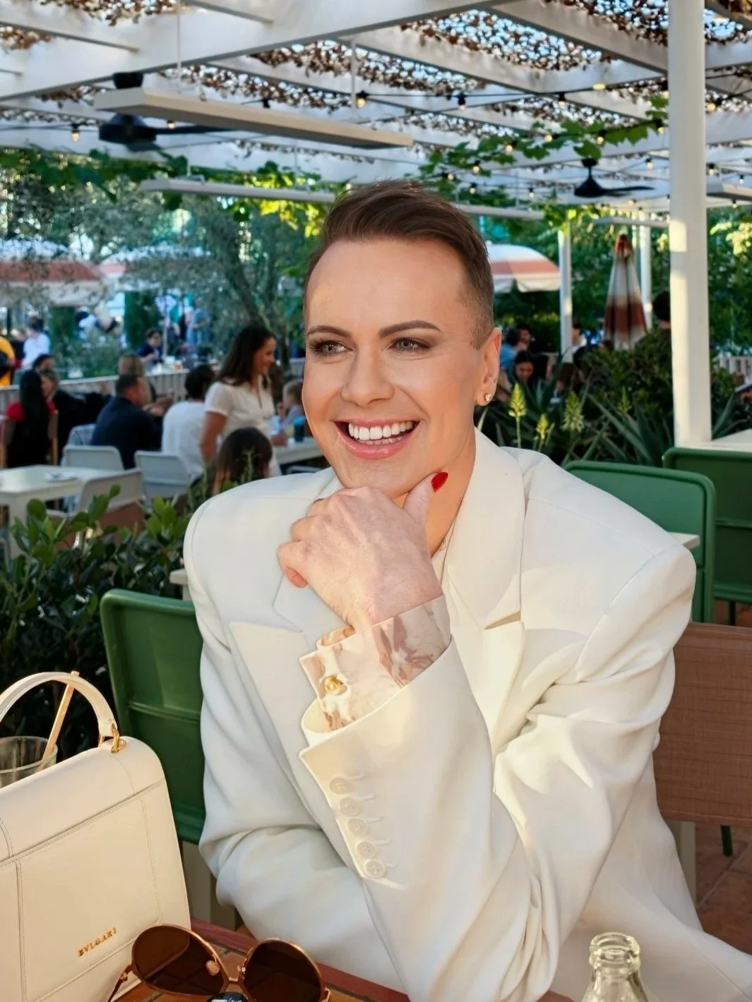 A smiling woman with short brown hair wearing a white blazer and floral shirt cuffs, sitting at an outdoor restaurant with green chairs and shaded by a pergola, surrounded by other diners and greenery.