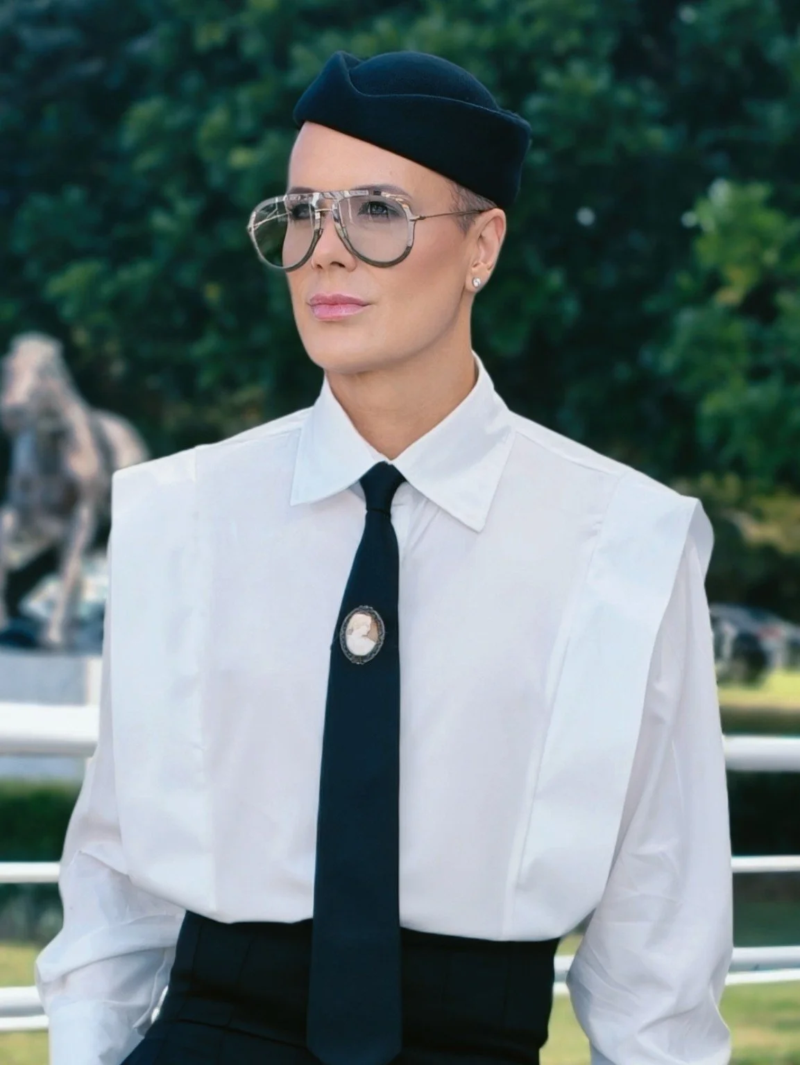A person, man, wearing a white shirt, black tie, and large glasses stands outdoors with green trees in the background.