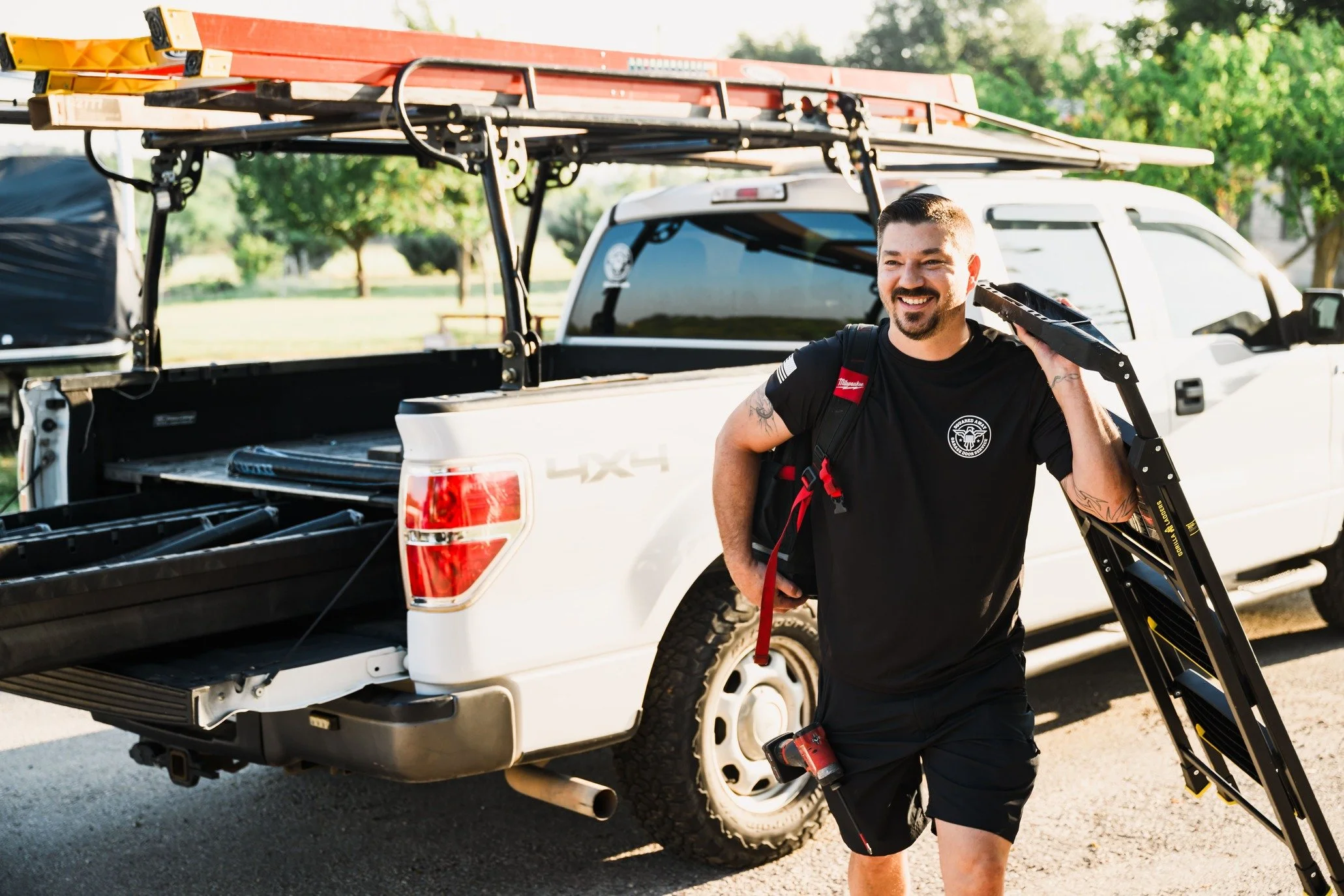 A smiling man walking away from a white pickup truck with surfboards and a ladder in the truck bed, holding a black step ladder, in a park with green trees.