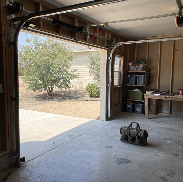 An open garage with a concrete floor, shelving and work table on the right, a duffel bag on the floor, and a view of the driv