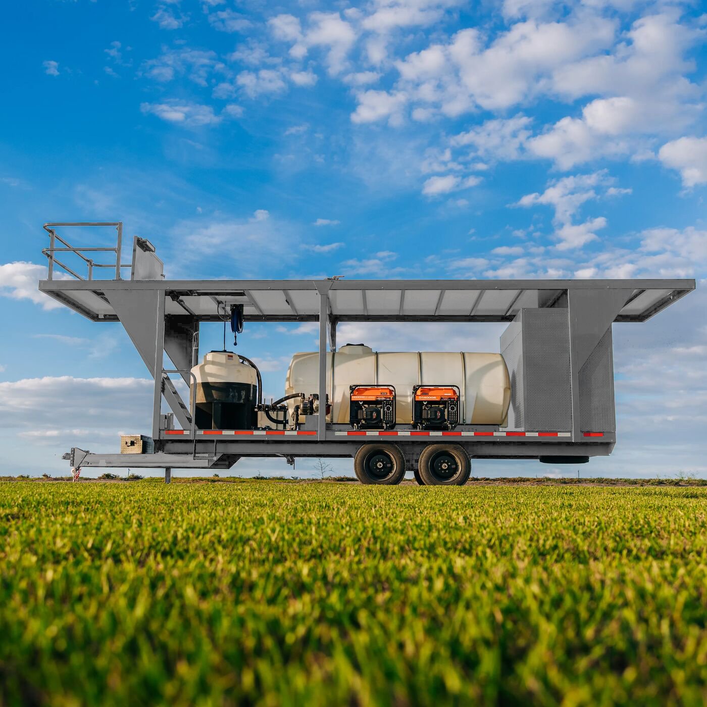 A large mobile water spraying machine with tanks and equipment, set on a grassy field under a bright blue sky with scattered clouds.