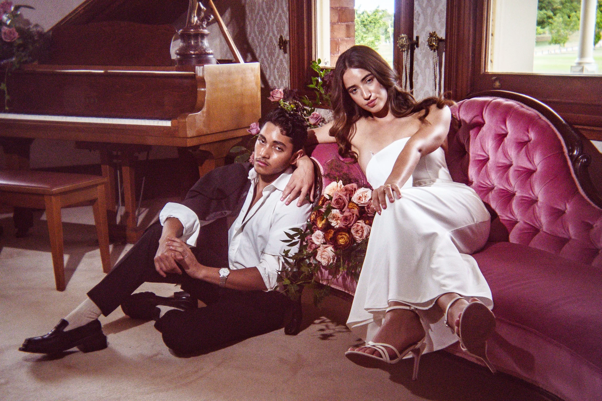 A young man and a young woman are together indoors, sitting on the floor near a pink velvet couch. The man is reclining with his head resting on the woman's lap and looking at the camera, while the woman is sitting upright, wearing a white dress, and holding a bouquet of pink roses. There is a wooden piano and large windows in the background.
