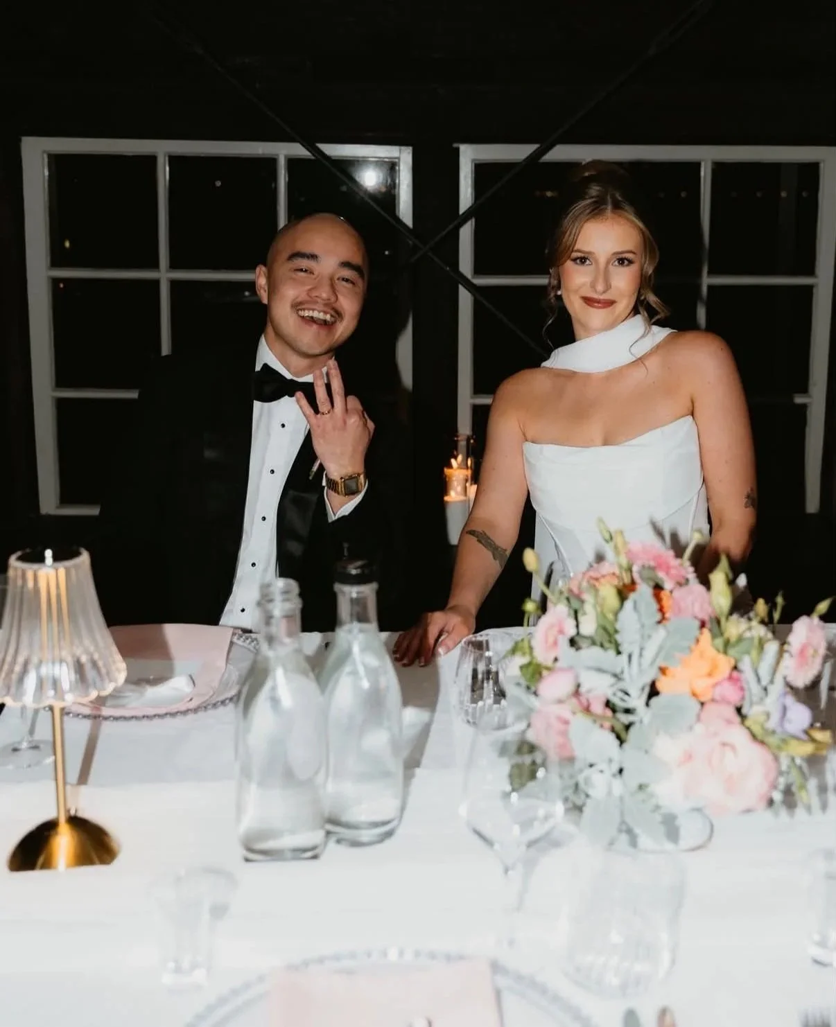 Bride and groom smiling together at their wedding reception, with a pastel floral centrepiece of pink, peach and white blooms on a candlelit table.