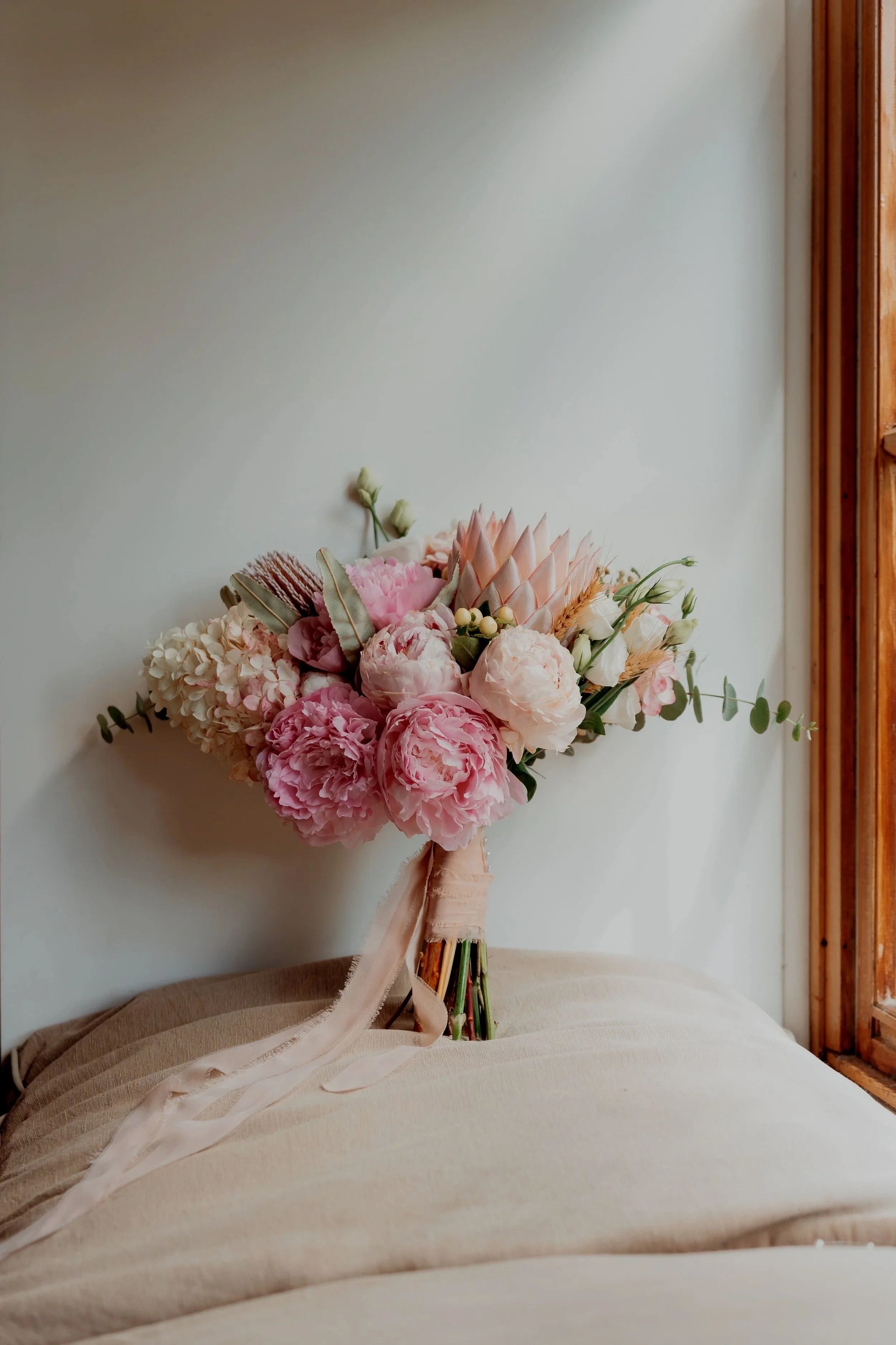 A bouquet of pink and white flowers, including hydrangeas and peonies, with greenery and a pink ribbon, resting on a beige surface near a white wall and a wooden window frame.
