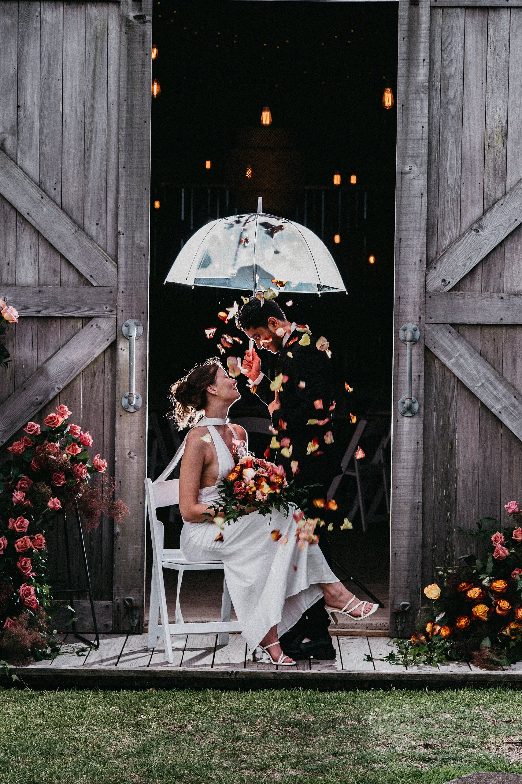 A couple at their wedding, sitting in a rustic barn doorway, with romantic lighting and flowers, sharing a moment surrounded by falling flower petals.