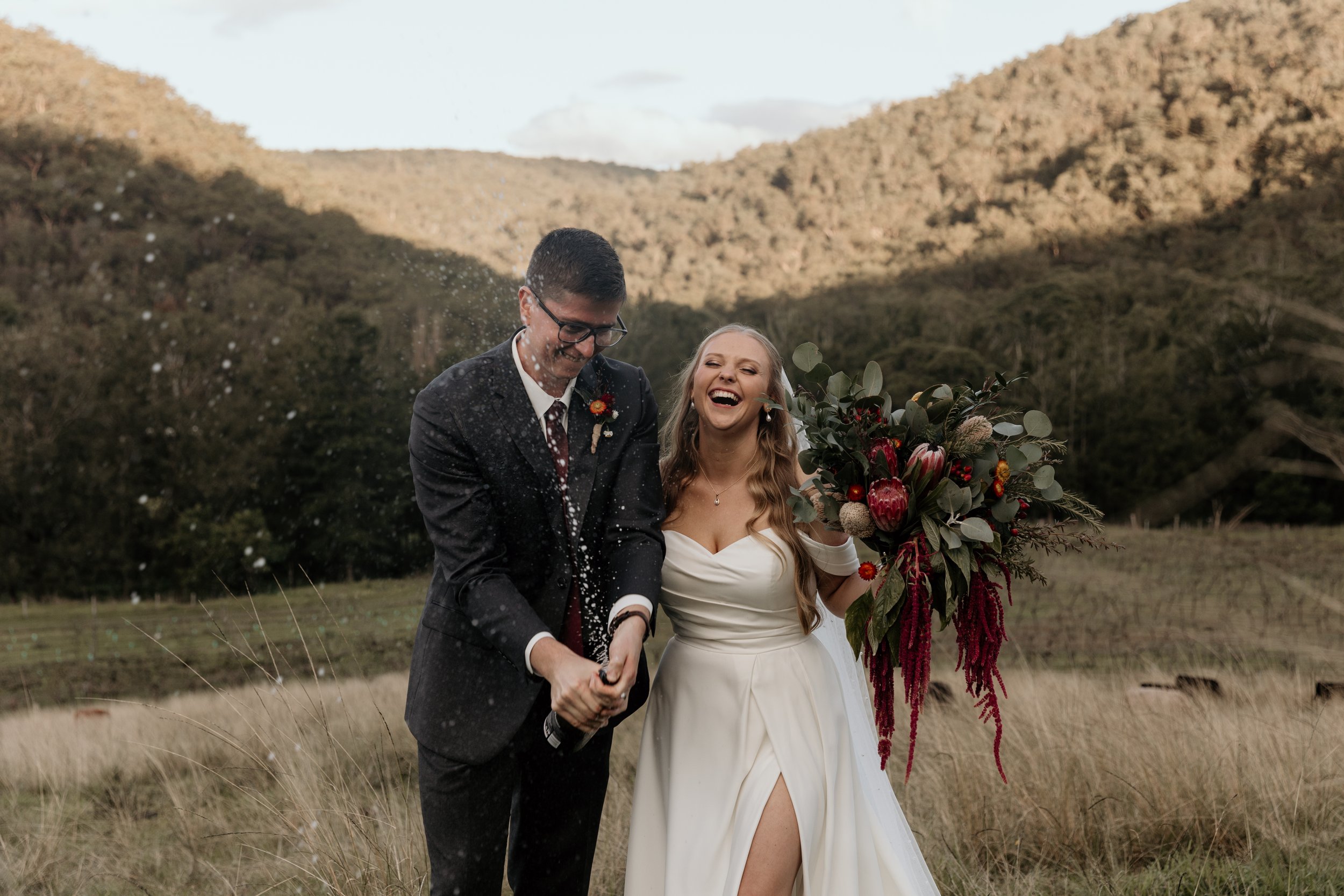 A bride and groom celebrating outdoors, holding hands, with the groom spraying champagne, and the bride smiling and holding a bouquet of flowers, in a rural landscape with mountains in the background.