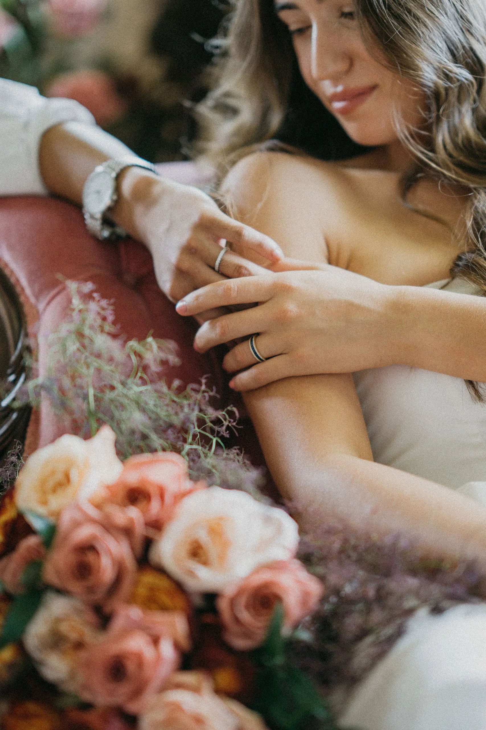 A woman with curly brown hair smiling with eyes closed, wearing a ring, lying in bed surrounded by pink and peach roses and greenery, with flowers and soft fabric in the foreground.