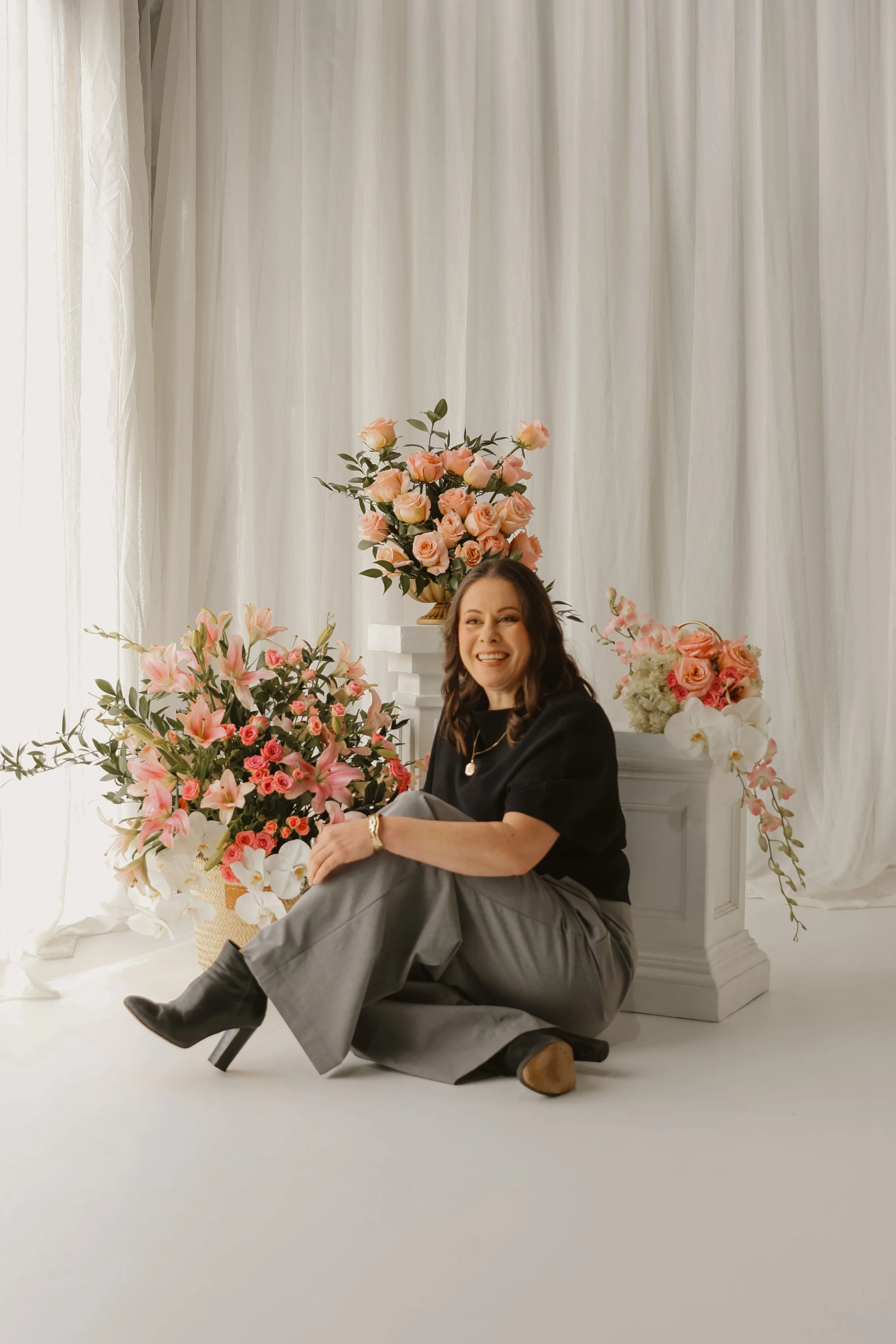 A woman sitting on the floor surrounded by large pink and white floral arrangements, smiling at the camera, with a white curtain backdrop.