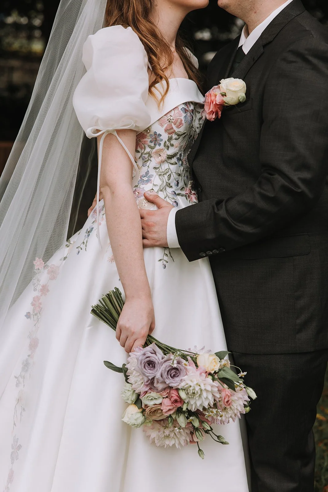 Close-up of a bride and groom holding each other at their wedding, with the bride holding a bouquet of pink, purple, and white flowers, and wearing a floral embroidered wedding dress with puffed sleeves.